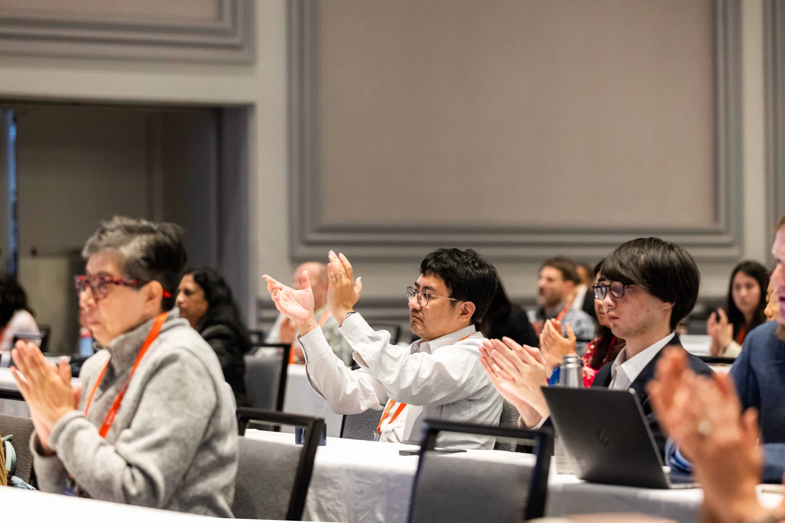 People attending a conference, sitting at tables, clapping.