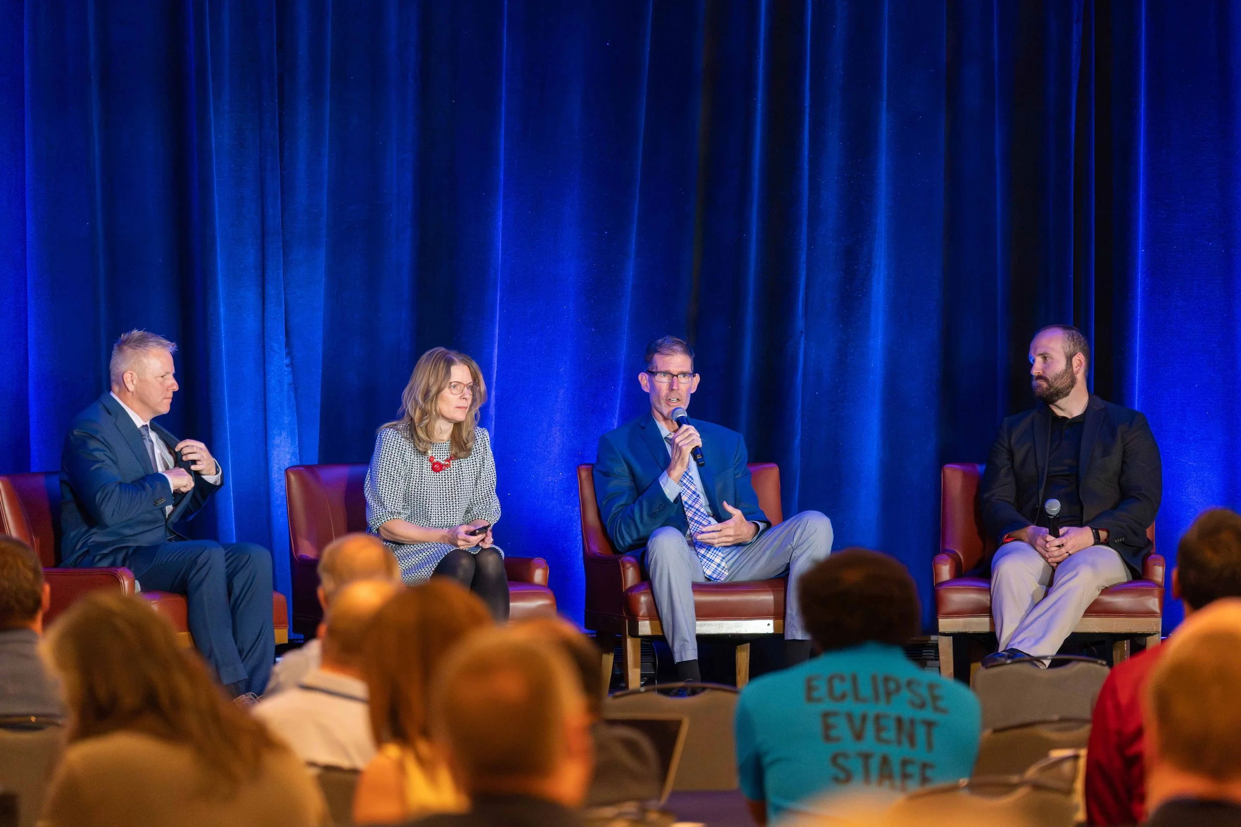 Panel discussion with four speakers on stage in front of a blue curtain, seated in leather chairs. Audience members visible in foreground, one wearing a shirt with 'ECLIPSE EVENT STAFF' on the back.
