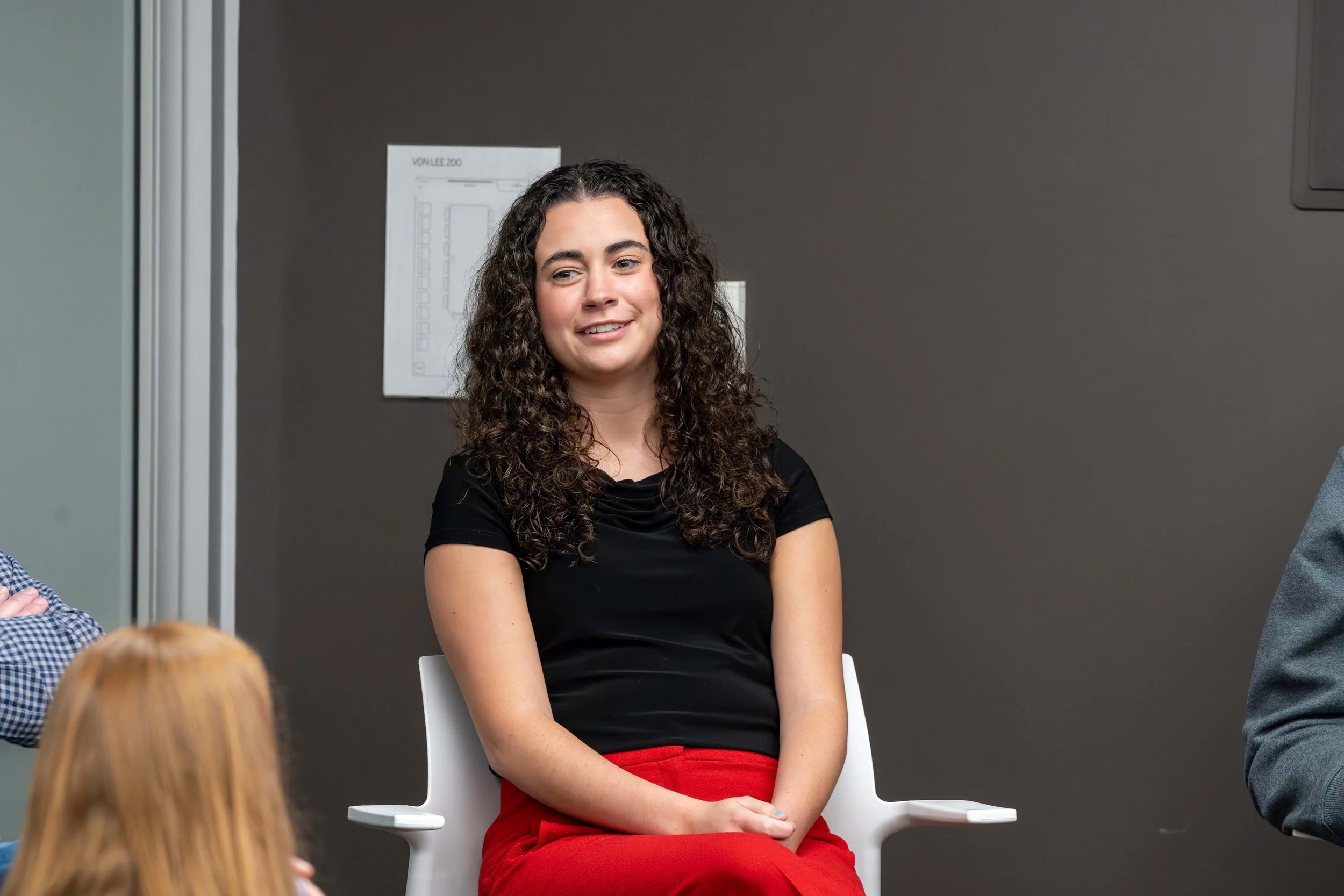 A young woman with curly dark hair wearing a black top and red pants, smiling and sitting in a white chair during a discussion or interview.