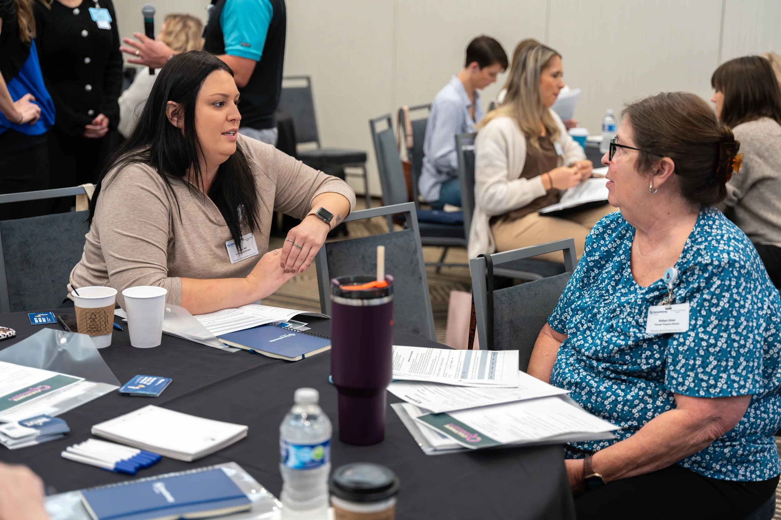 Two women engaged in conversation at a conference table with notebooks, papers, a water bottle, and disposable cups in a meeting room. Other attendees are seated and standing in the background.