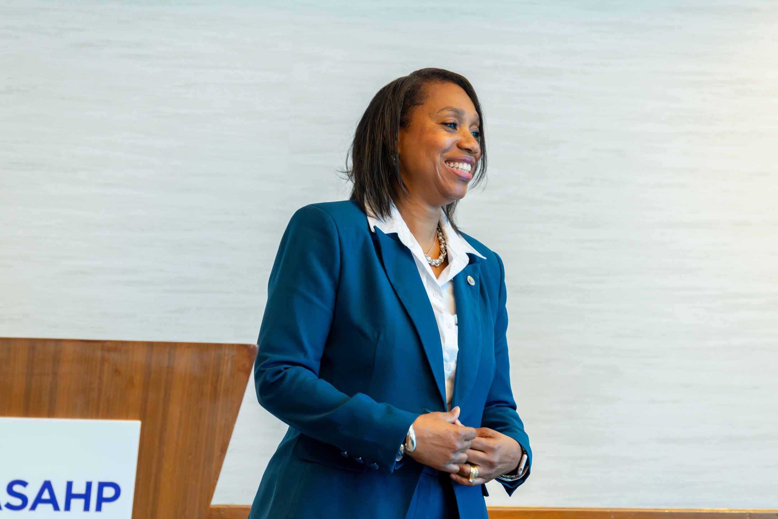 A woman in a blue blazer and white shirt smiling during a presentation or speech in a professional setting.