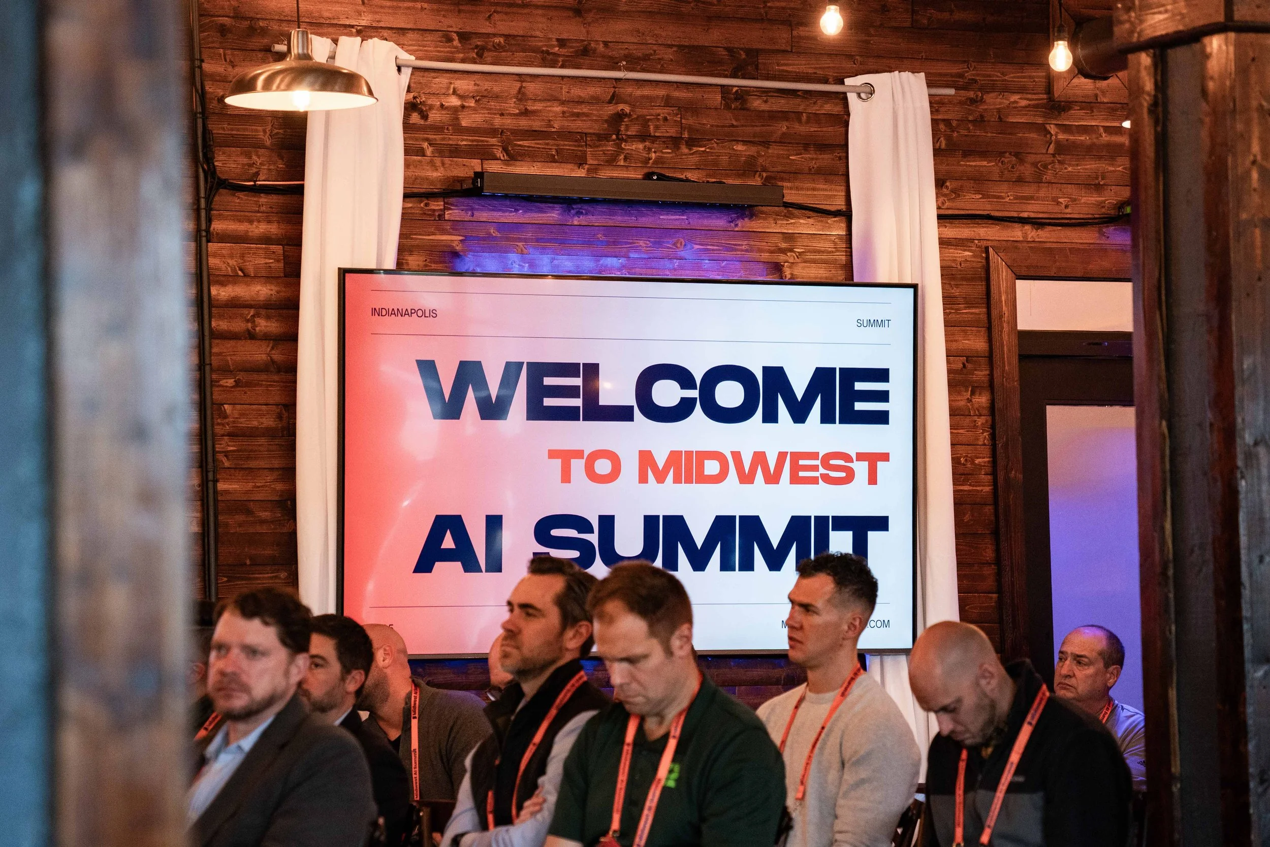 A large screen displaying 'Welcome to Midwest AI Summit' with attendees seated in front, inside a room with wooden walls and white curtains.