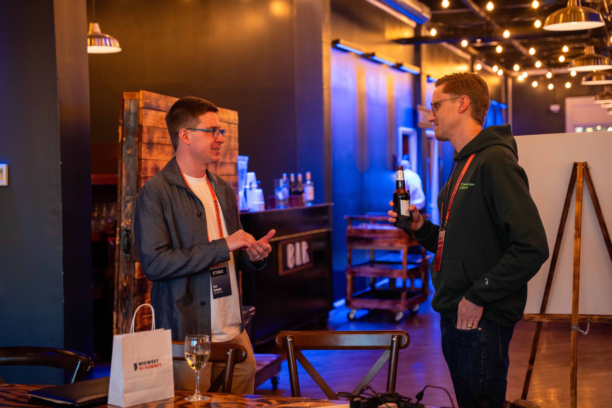 Two men having a conversation at a bar or conference event, one holding a beer bottle, in a dimly lit room with blue and yellow lighting, wooden furniture, and a bar in the background.
