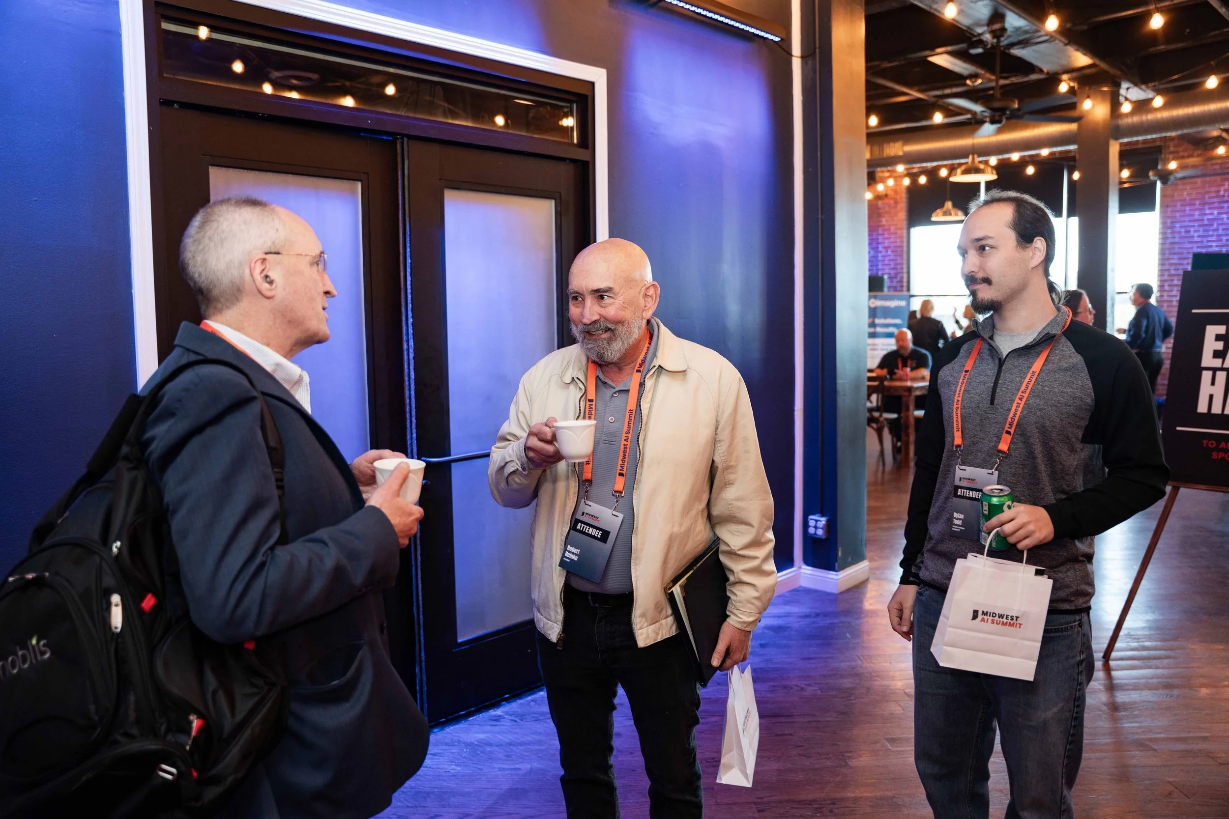 Three men engaged in conversation at a conference. Two of them are holding coffee cups, one is holding a paper bag and a brochure. They are wearing conference badges and lanyards, standing inside a venue with warm lighting, brick walls, and visible c