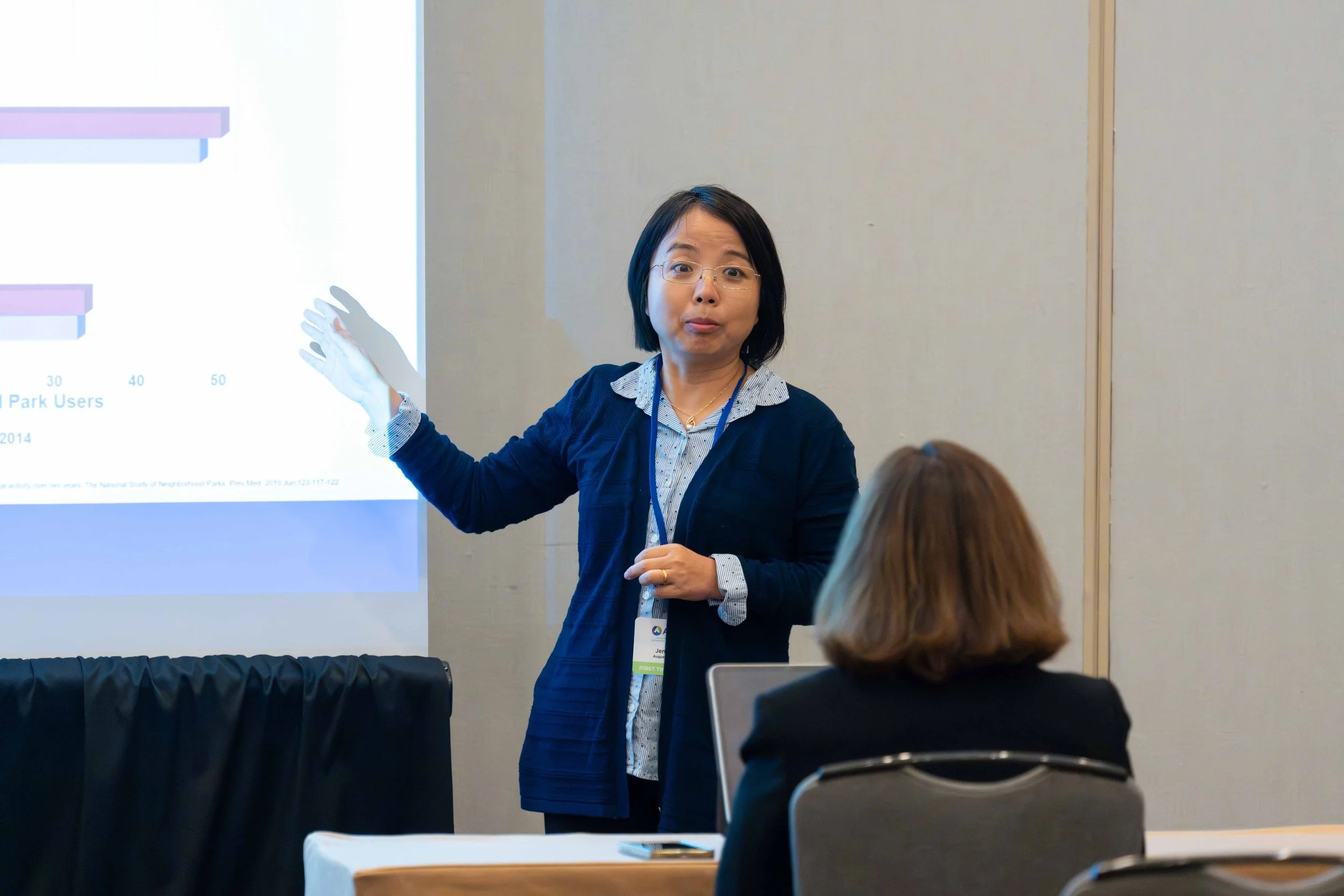 A woman with glasses and a blue cardigan gives a presentation to a seated audience member in a conference room.