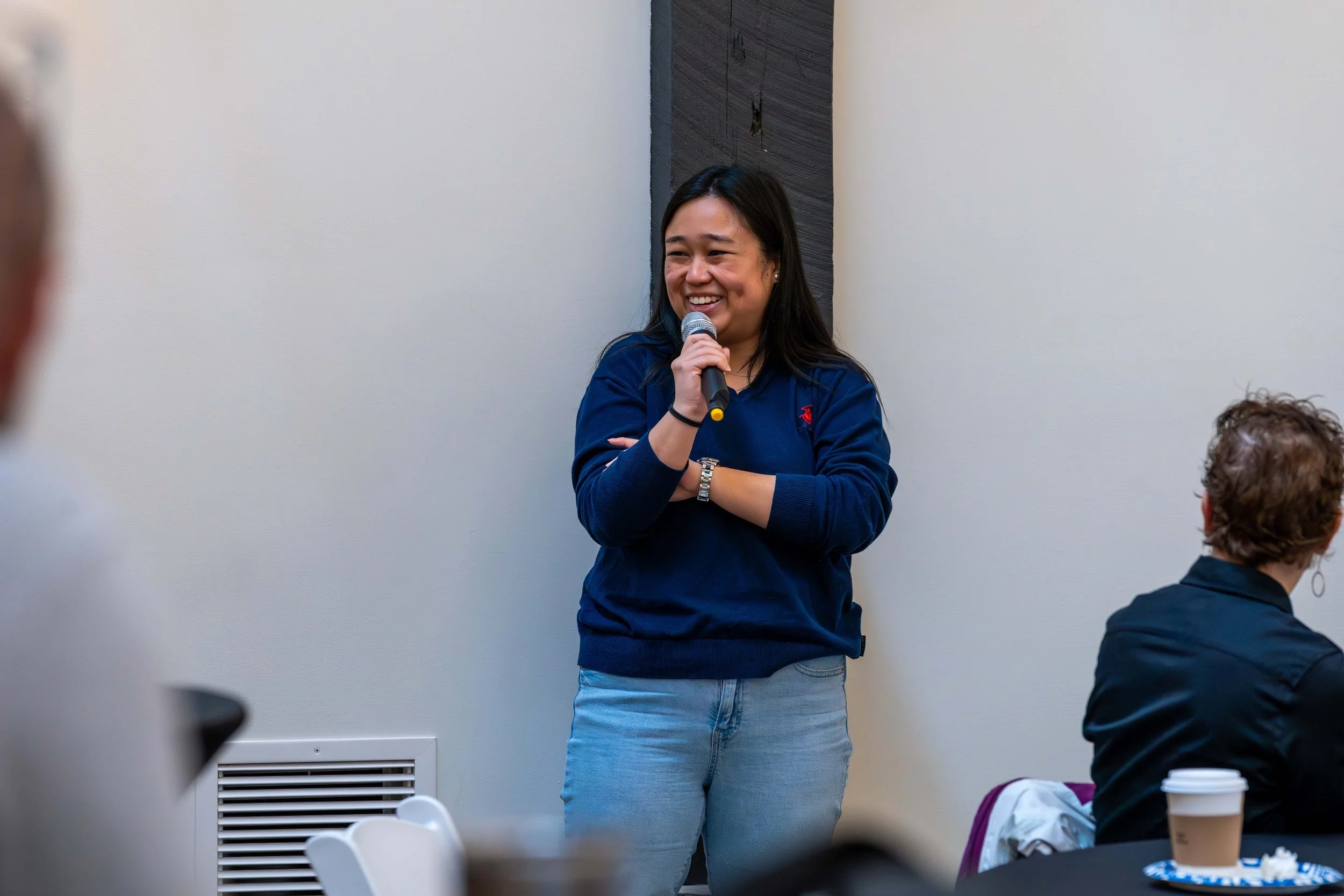 A woman in a blue sweater holding a microphone and smiling during a speech or presentation at a meeting or event.