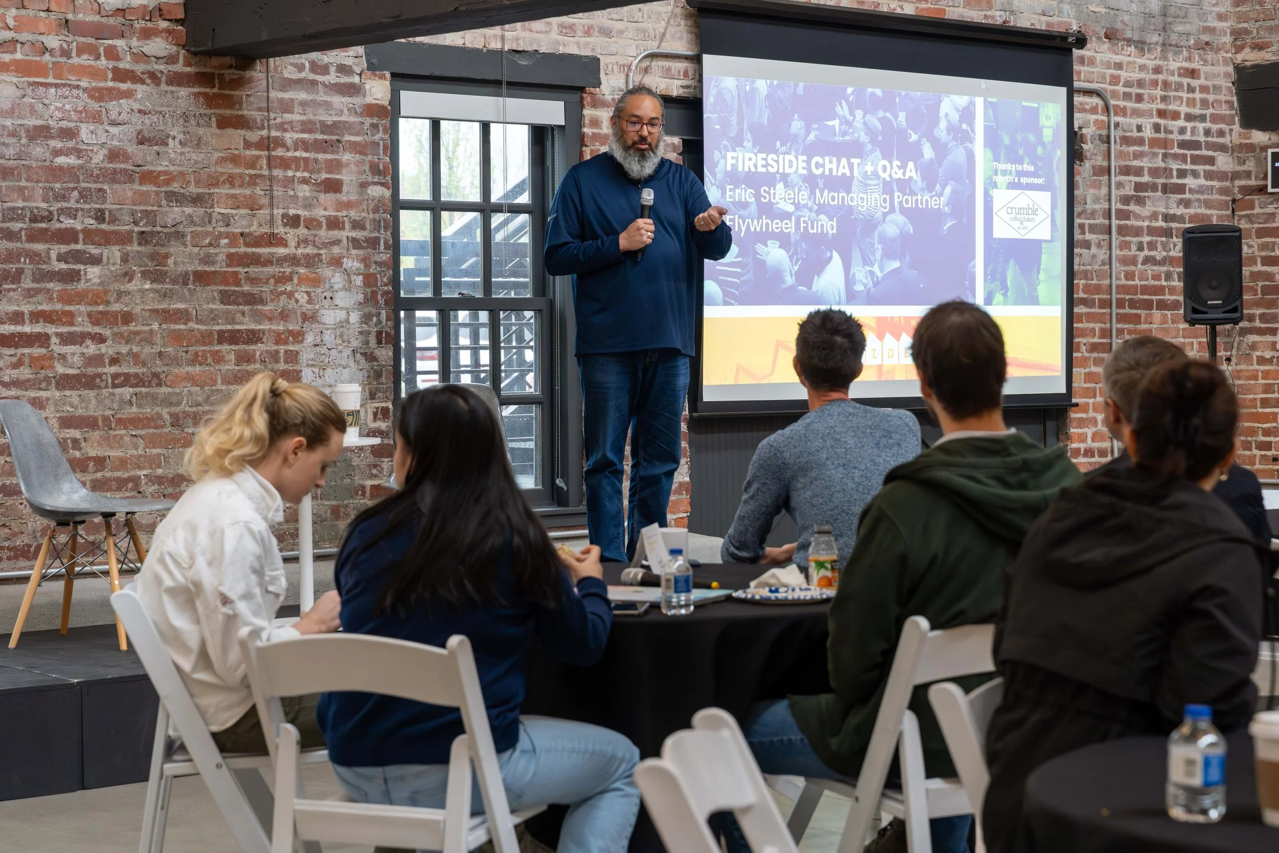 A man giving a presentation on stage with a microphone in front of a screen that reads 'Fireside Chat + Q&A' and includes his name 'Eric Steele, Managing Partner, Flywheel Fund.' Several people are seated at round tables with black tablecloths, facin