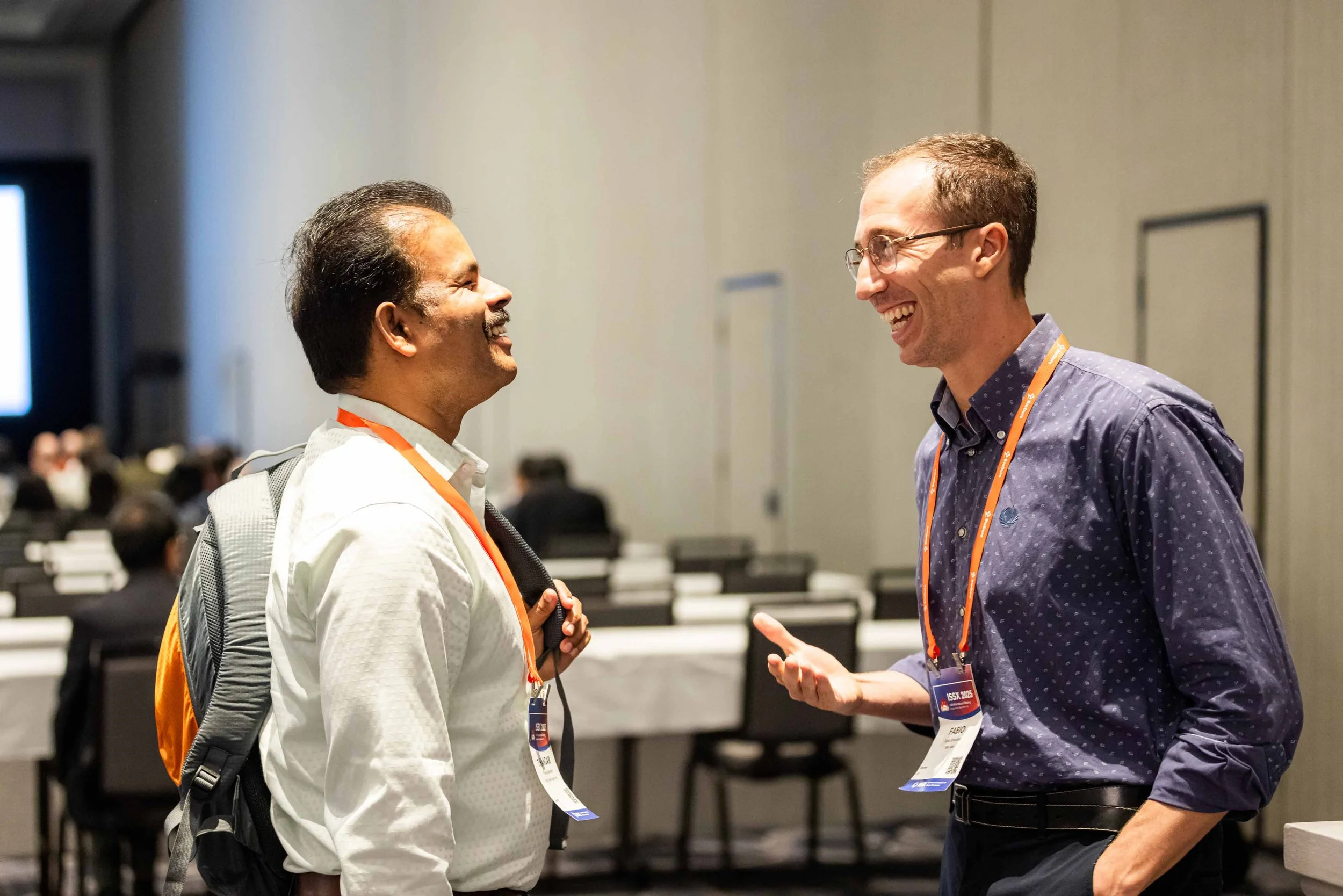 Two men are smiling and engaged in conversation at a conference or seminar, wearing conference badges and lanyards.