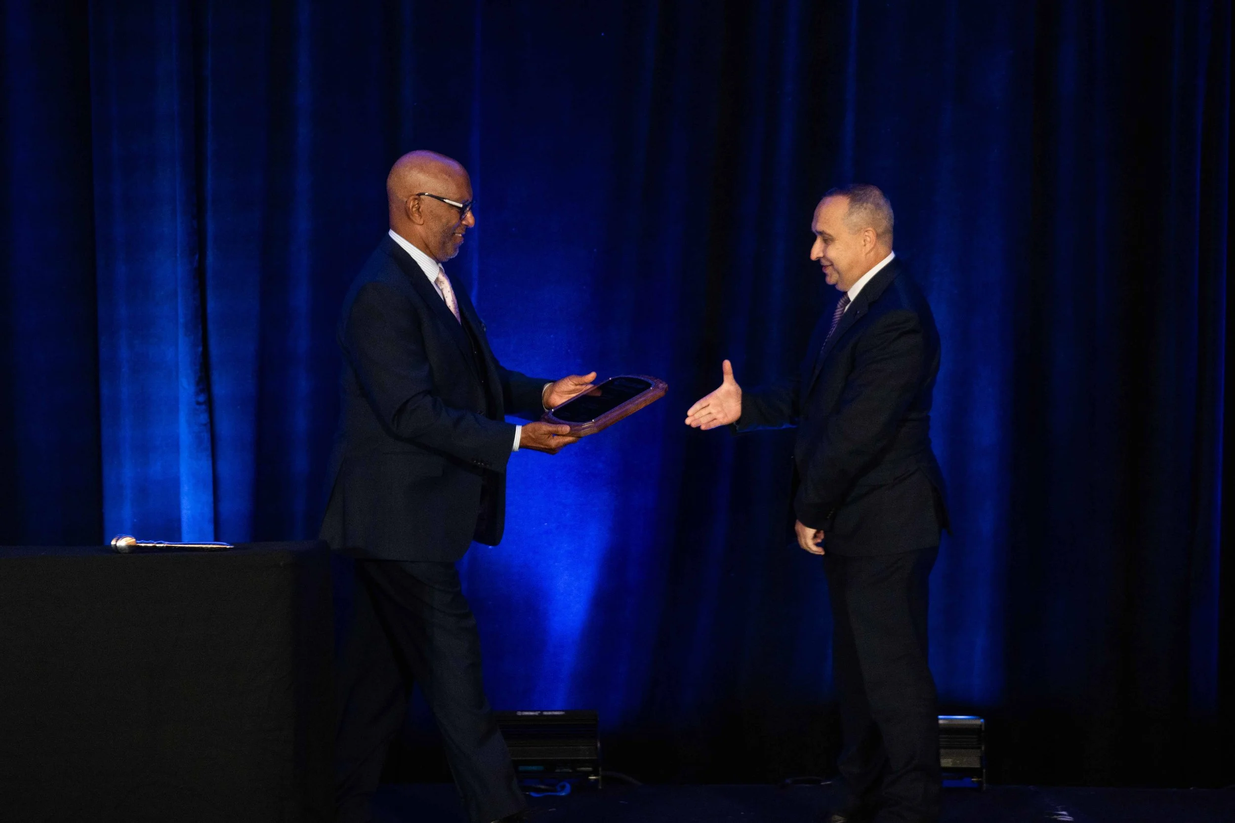 Two men in suits on a stage with a dark blue curtain background; one presenting an award or plaque to the other.