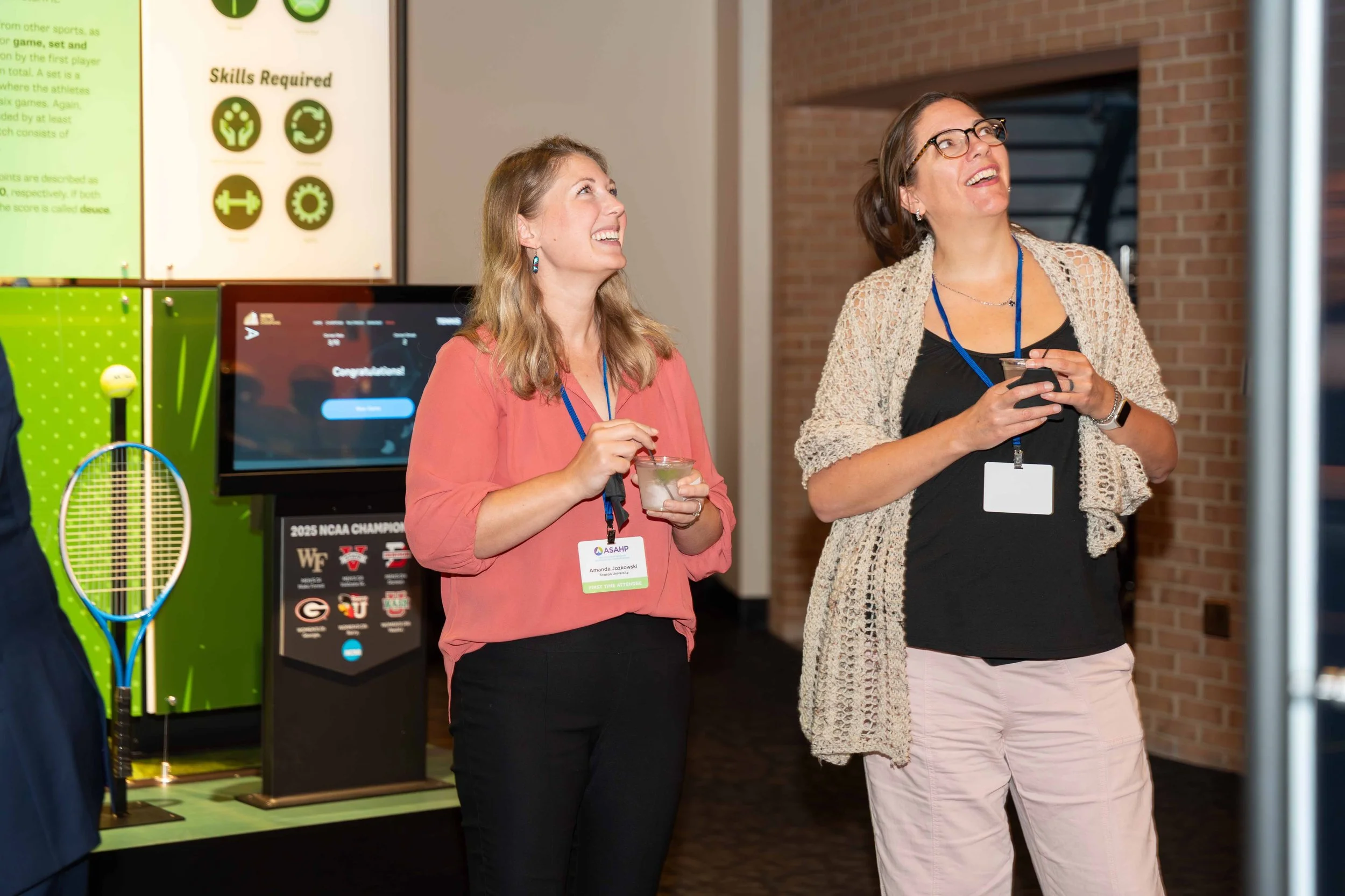Two women smiling and looking upward at an indoor event, one holding a drink and the other holding a phone, with conference badges around their necks. In the background are a green memorabilia display, a computer screen, and a brick wall.