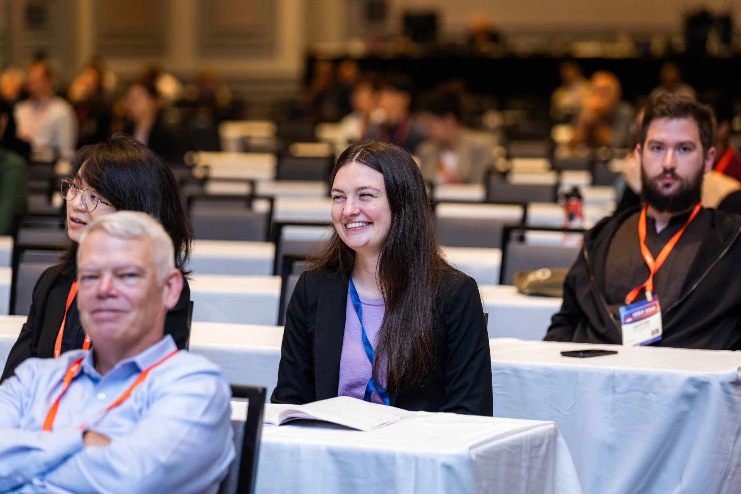 Group of people sitting at tables in a conference or seminar, smiling and paying attention to a presentation