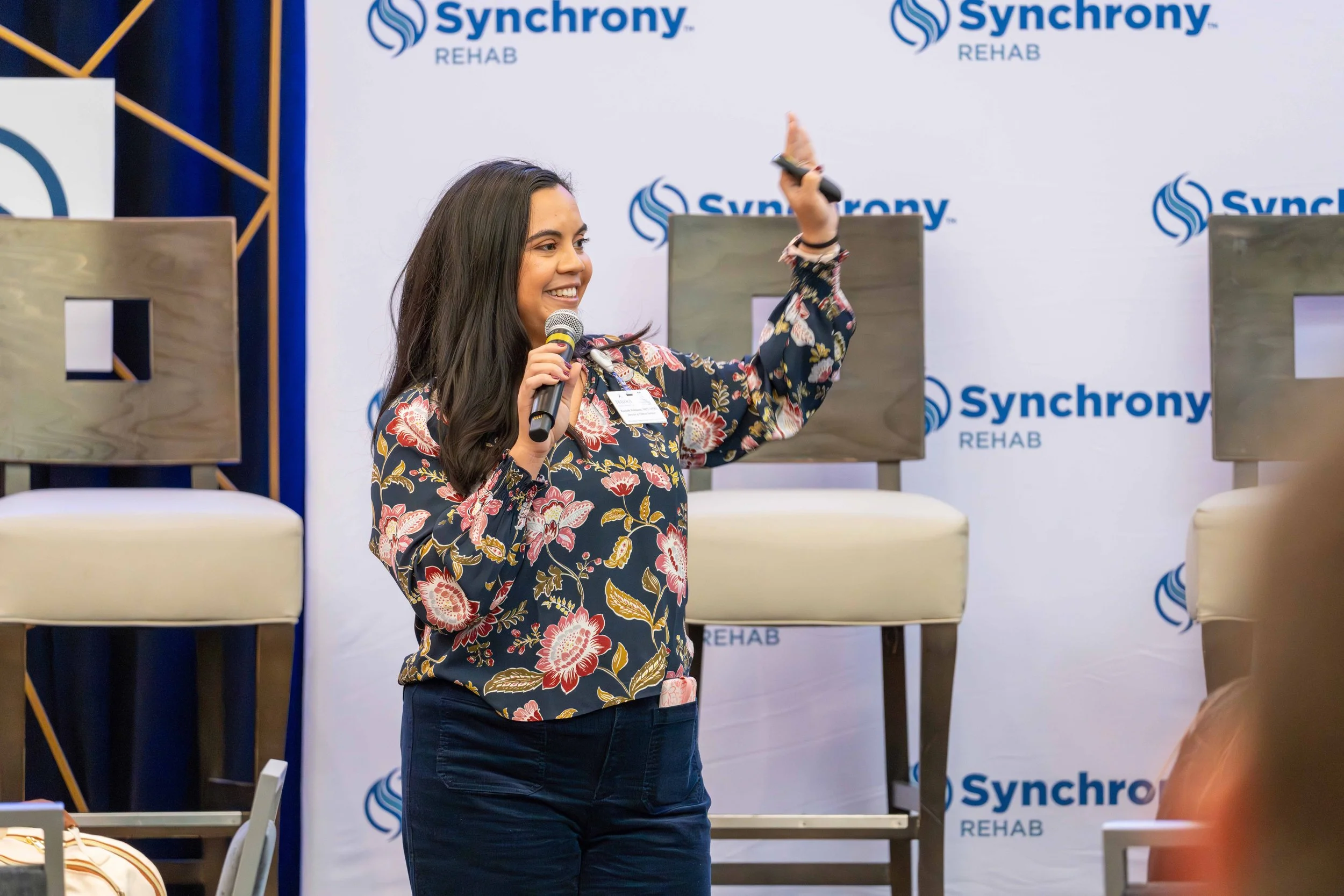 A woman with long dark hair holding a microphone and gesturing with her other hand, speaking at an event in front of a backdrop with the logo 'Synchrony Rehab'. She is wearing a floral blouse and dark pants, standing on a stage with chairs behind her