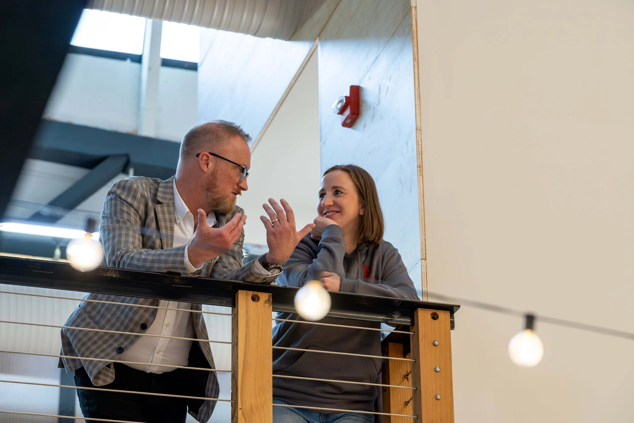Two people having a conversation on a balcony inside a modern building with string lights and a wooden railing.