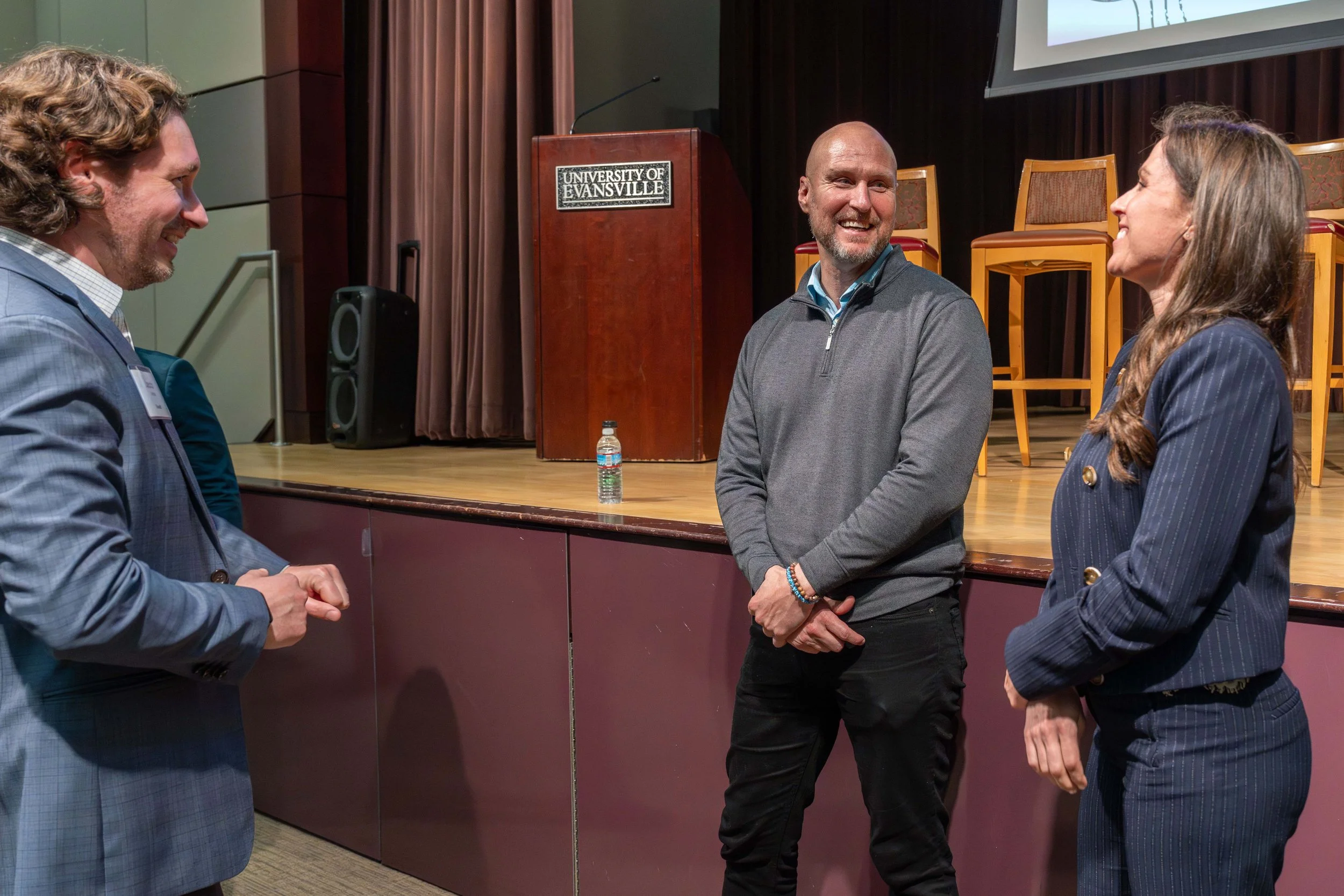 Three people having a conversation at an event at the University of Evansville. One man is wearing a blue plaid suit, a woman is wearing a navy pinstripe blazer, and another man in a gray zip-up sweater.