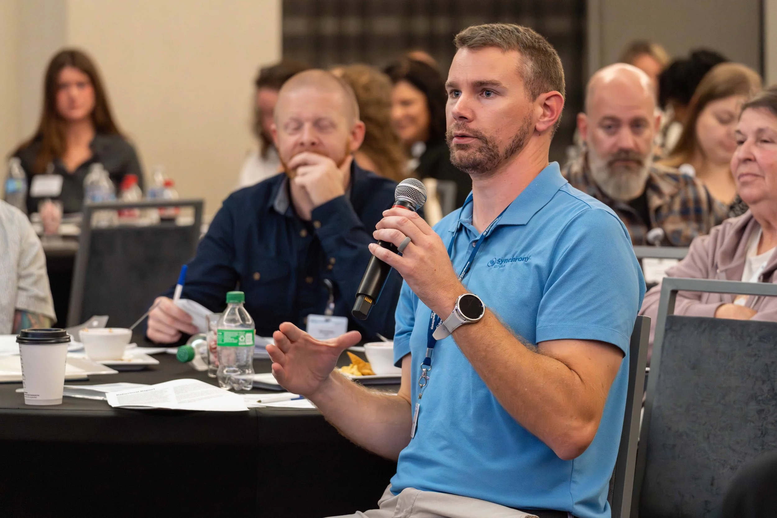A man in a blue polo shirt holding a microphone and speaking at a conference with several attendees listening attentively.