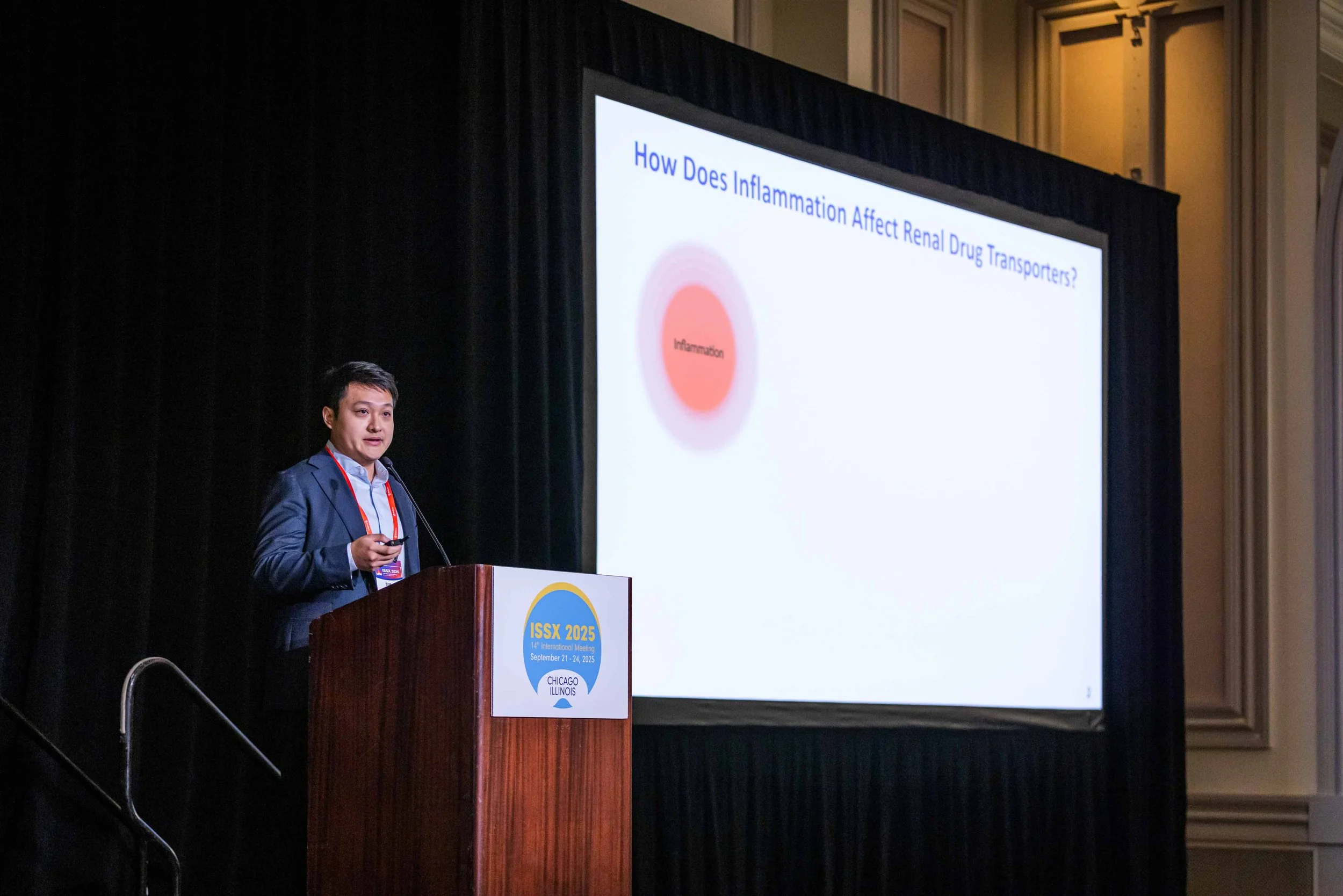 A man in a dark suit is giving a presentation at a conference, standing behind a wooden podium with a sign that reads 'ISSS 2025, 14th International Meeting, September 21-24, 2025, Chicago Illinois'. He is holding a remote control and speaking to the