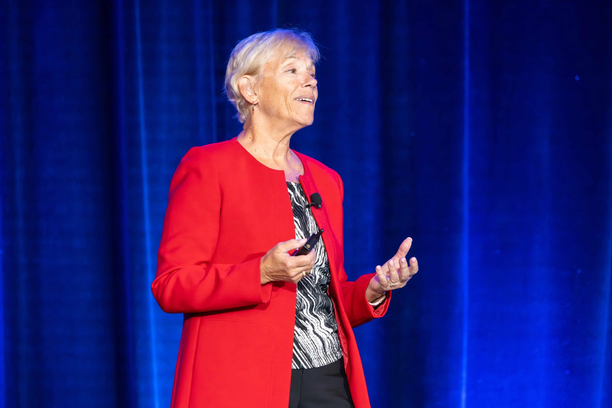 A woman with short gray hair wearing a red blazer and patterned blouse, speaking on a stage with a blue curtain background.