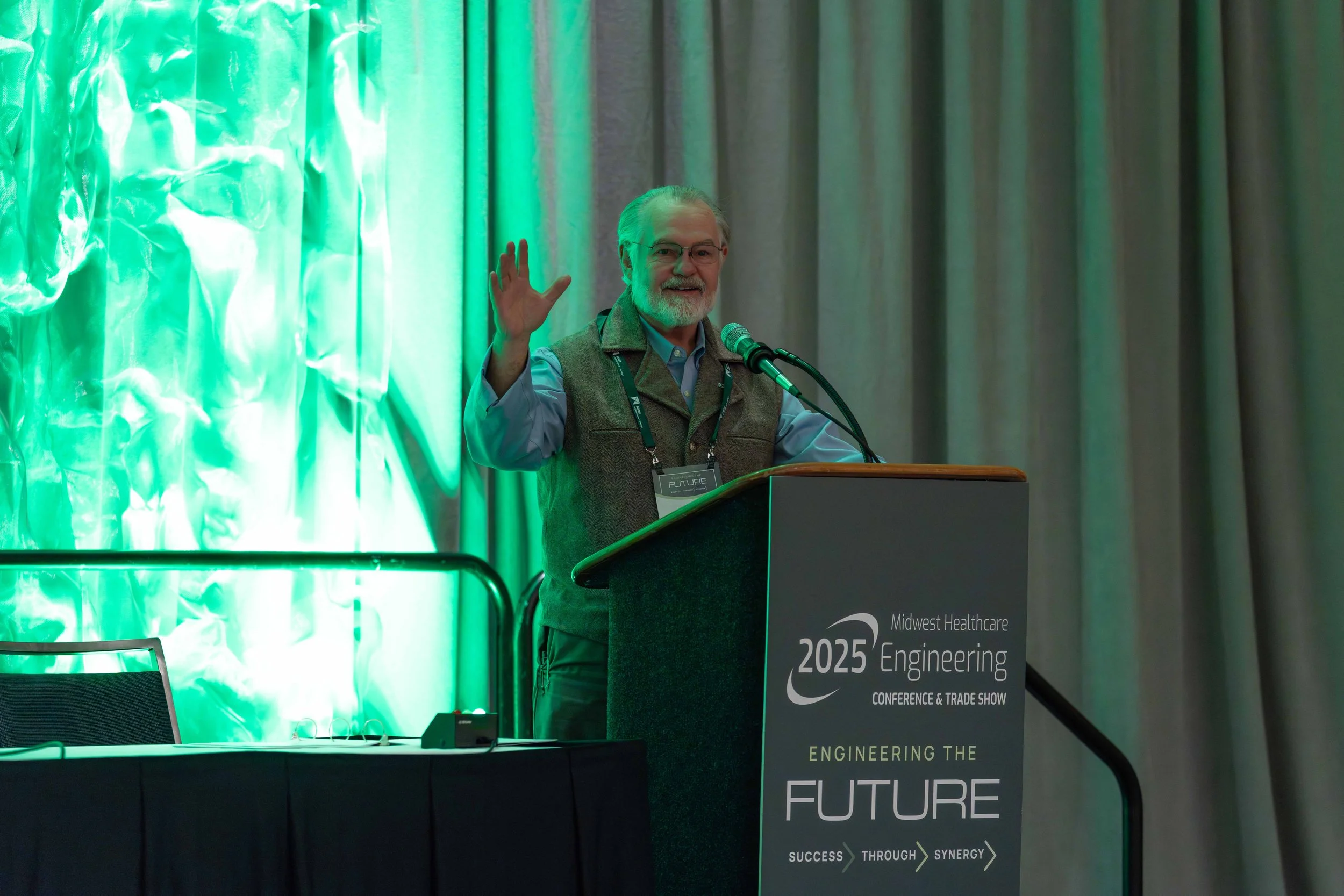 Man speaking at a podium during Midwest Healthcare Engineering Conference 2023, with a backdrop of green lighting and curtains.