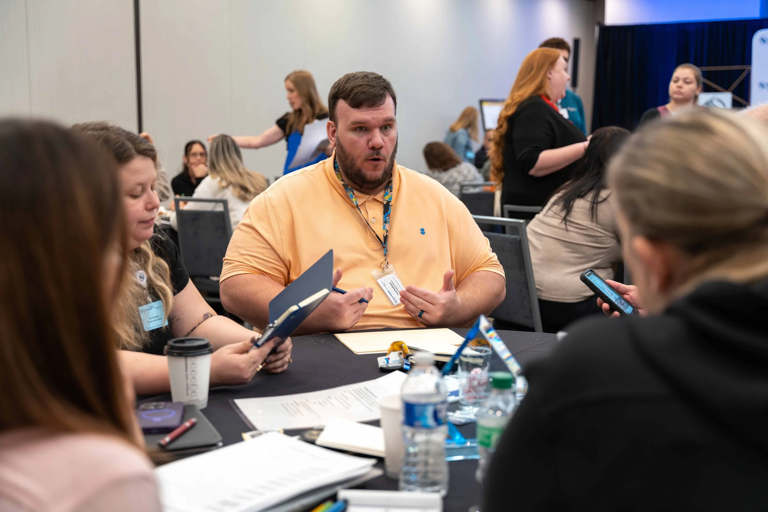 Man in peach shirt and badge engaging in conversation at a conference table with women, some holding phones or notebooks, in a busy conference room.