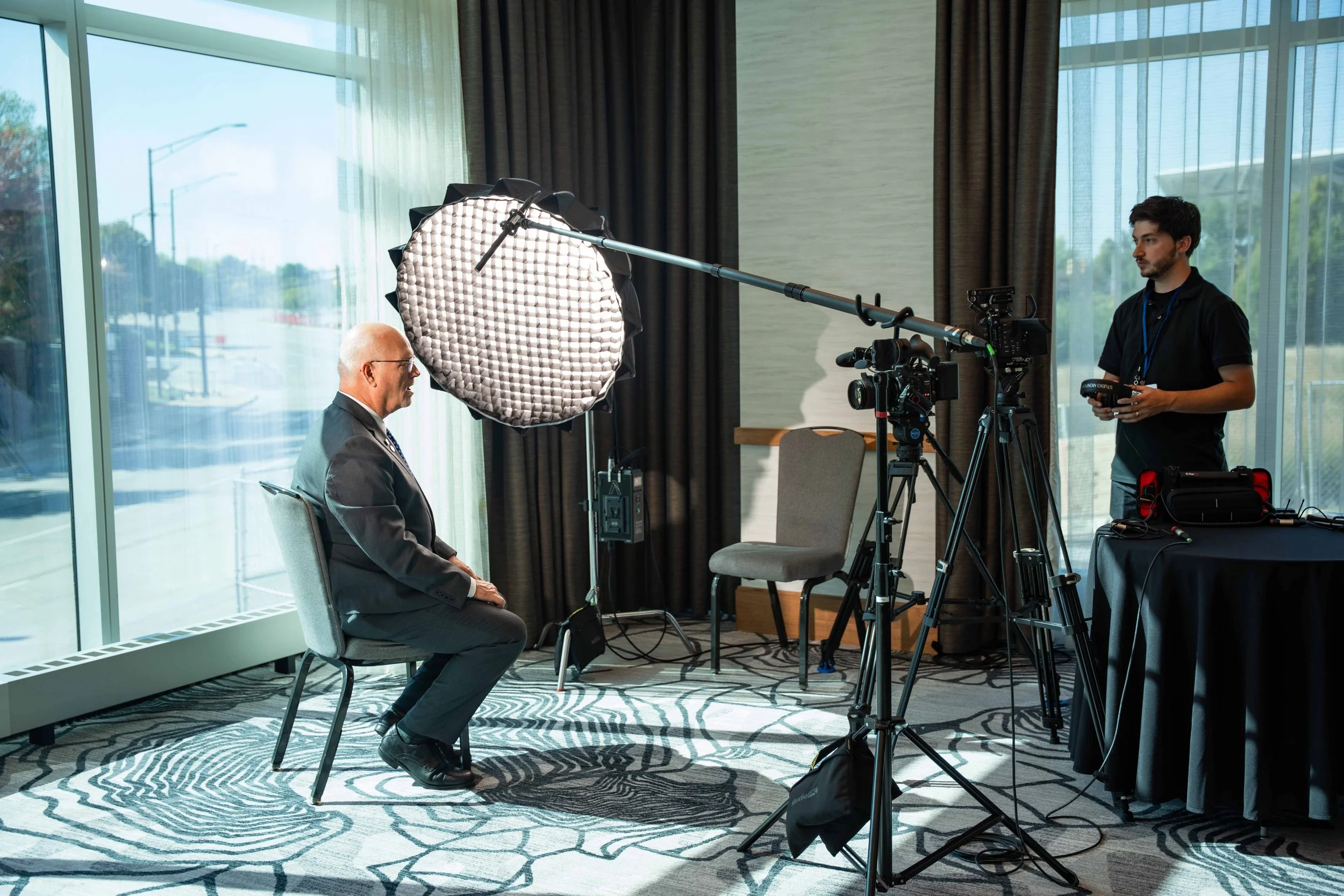 A man in a suit is being filmed or photographed during an interview or recording session in a bright room with large windows. A person on the right is operating camera equipment and holding a remote control.