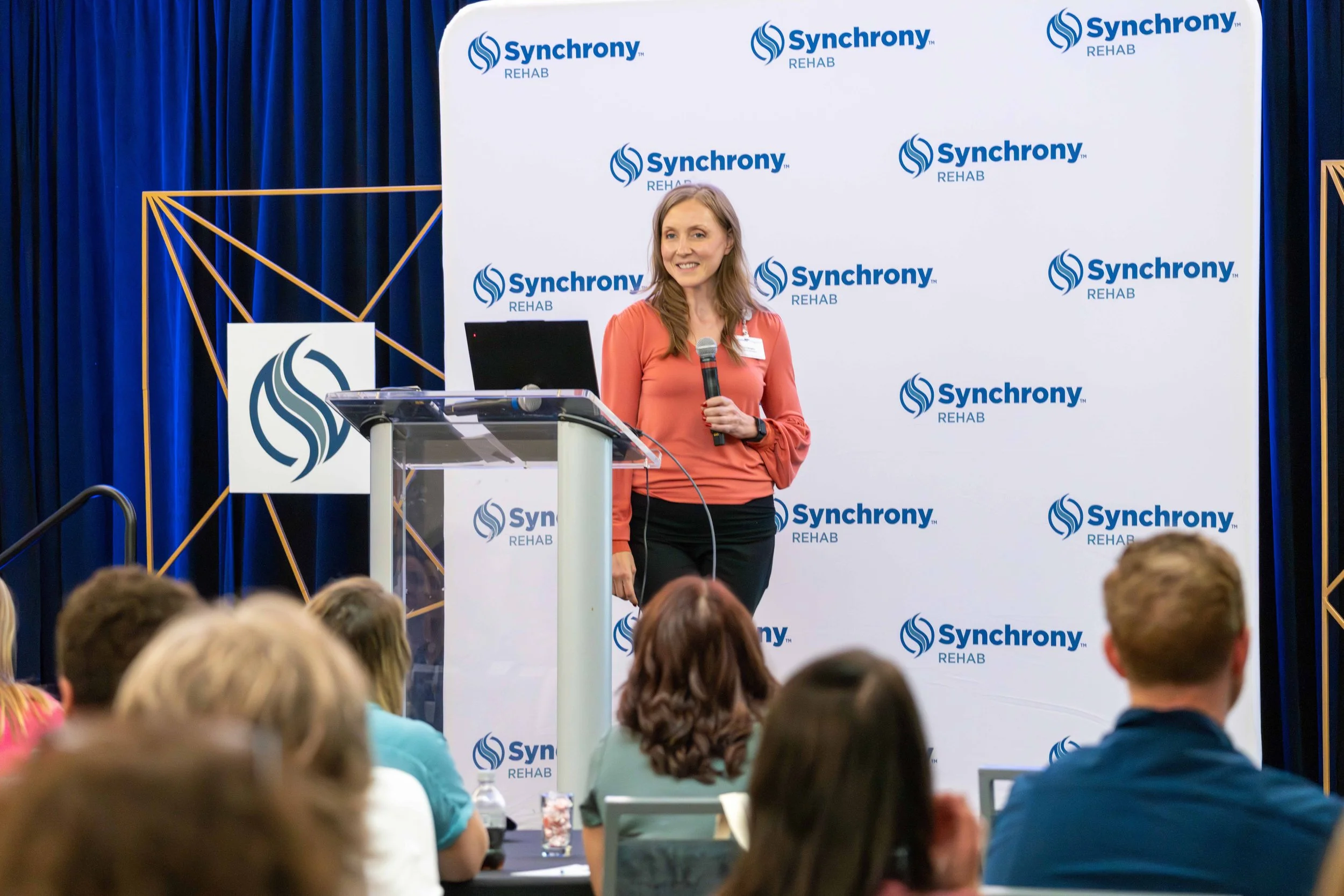 A woman speaking at a conference on stage with a backdrop featuring the 'Synchrony Rehab' logo. She is holding a microphone and standing next to a podium, with an audience seated in front of her.
