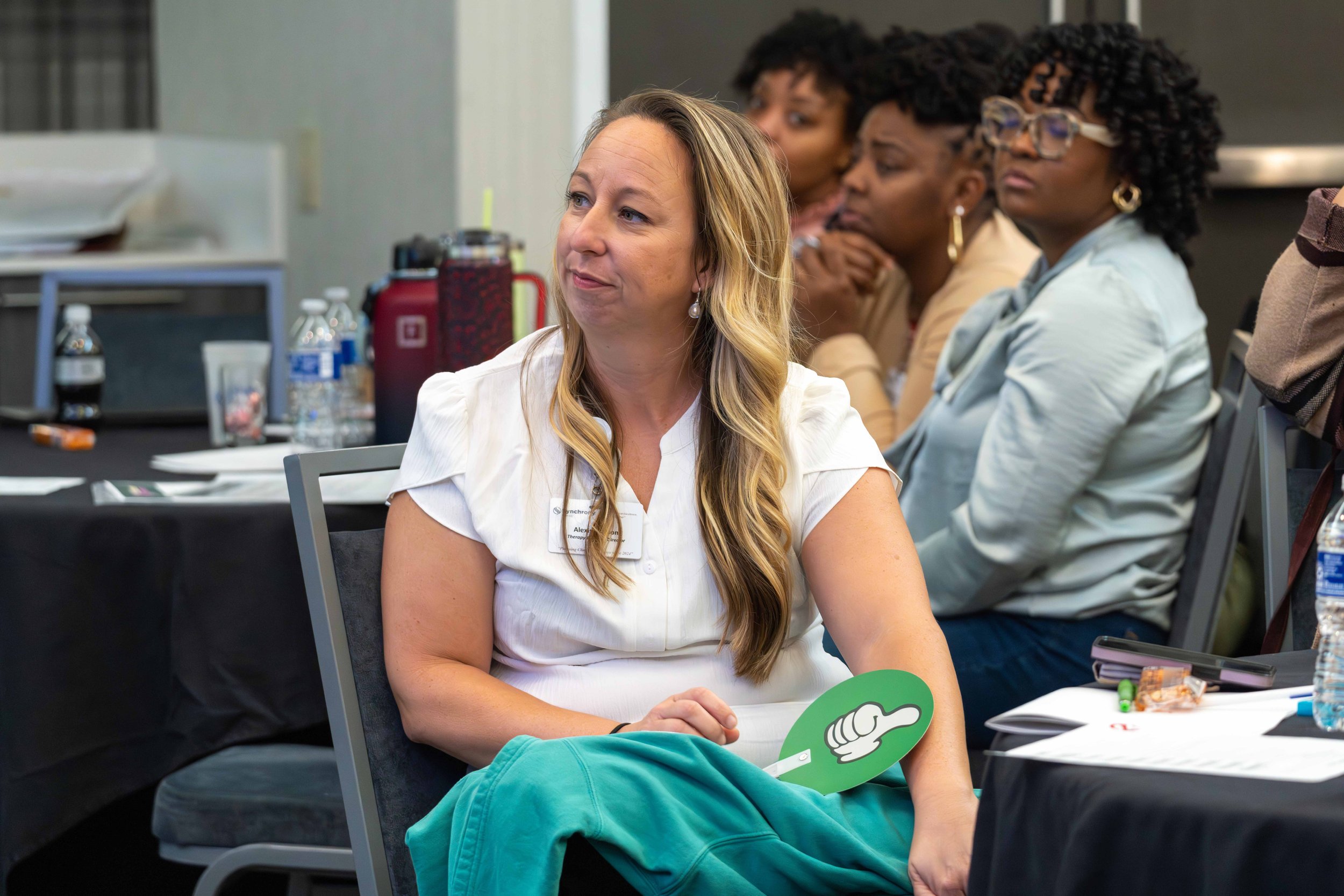 A woman with long hair sitting in a conference, holding a green sign with a thumbs-up symbol, surrounded by other women and conference materials on a table.