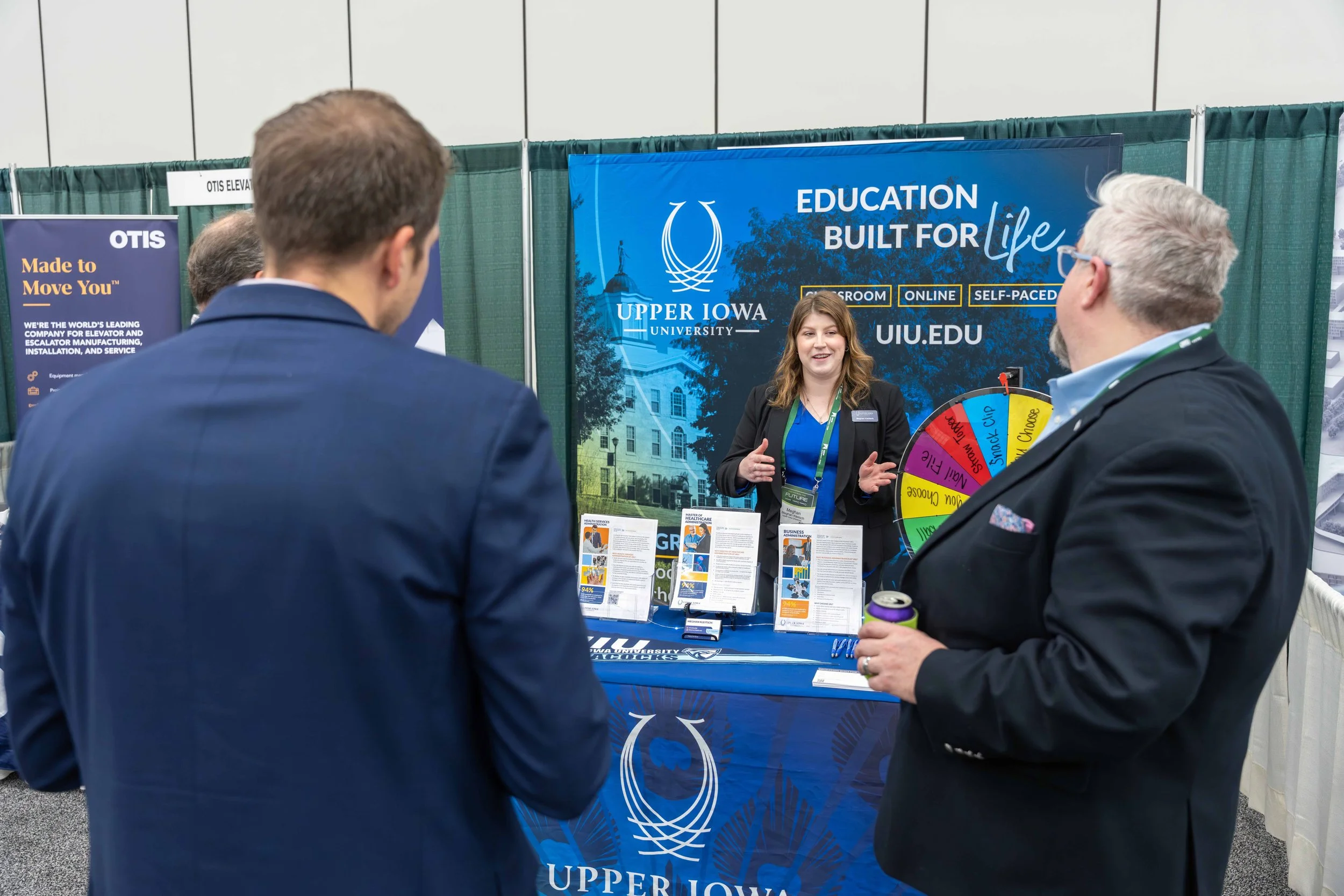 At a college fair, a woman in a black blazer is speaking to two men in suits at the Upper Iowa University booth, which features a large blue banner and a spinning wheel game.