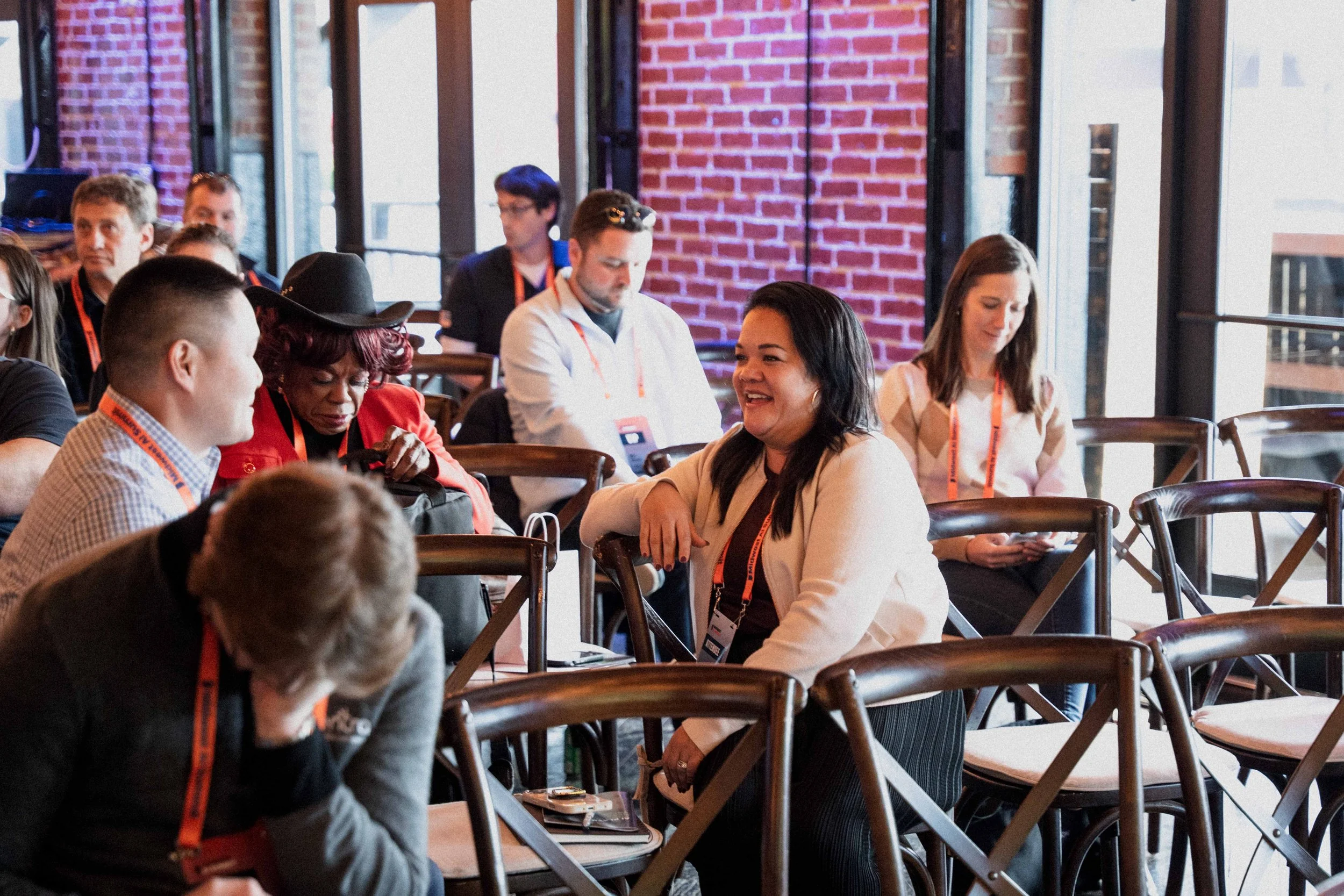 People sitting at tables in a conference room with exposed brick walls, engaging in conversation and using laptops, during a professional event.