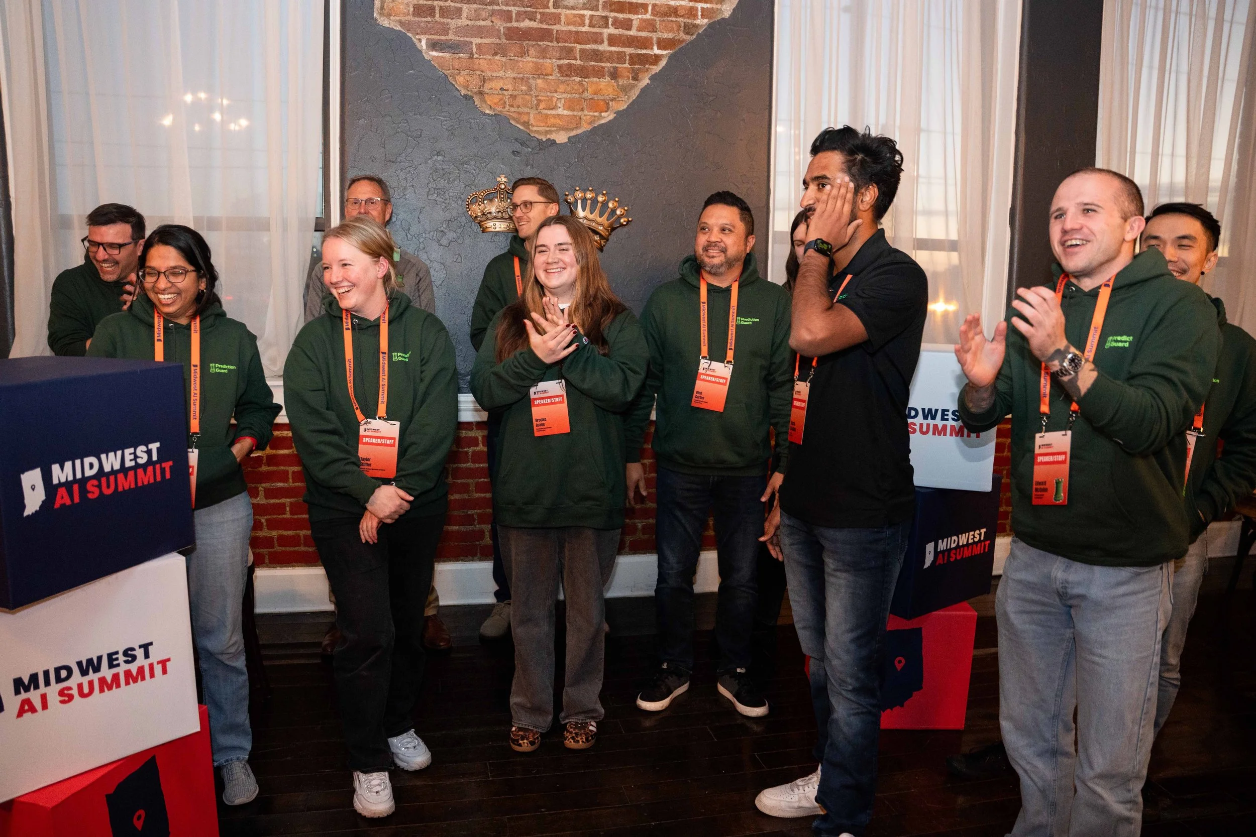 Group of smiling people at the Midwest AI Summit, some wearing green hoodies with event badges, standing in a room with brick and textured wall background, and a sign reading 'MIDWEST AI SUMMIT' visible.