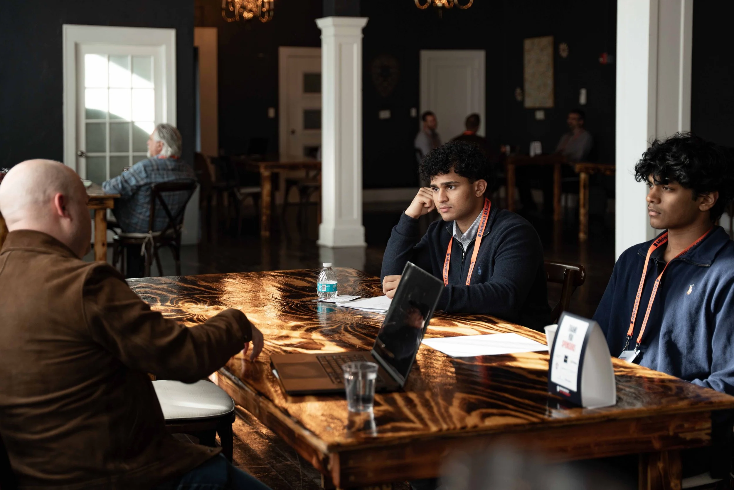 Two young men and an older man sitting at a large wooden table engaged in conversation in a well-lit room with dark walls, white trim, and some people in the background.