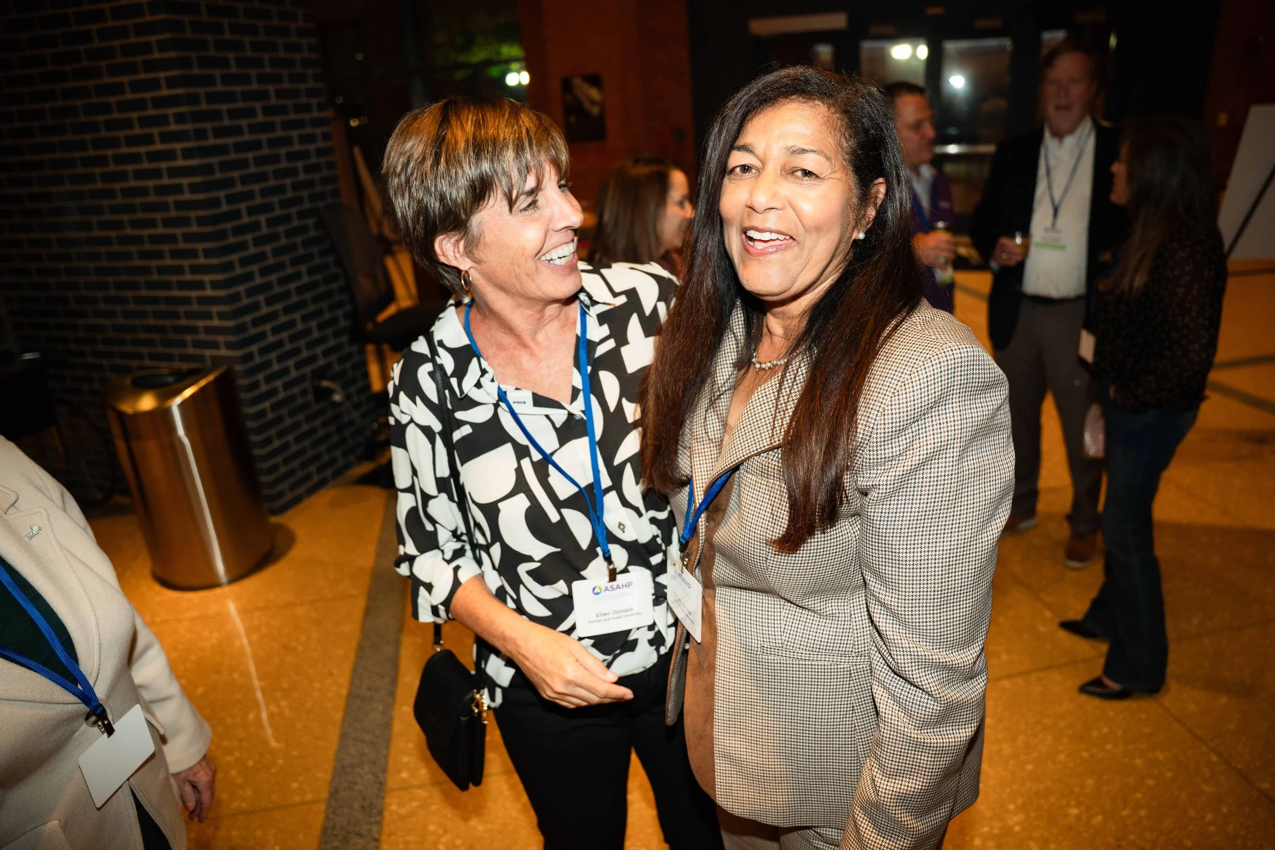 Two women laughing and talking at a professional event or conference, wearing name badges, with several other people in the background.
