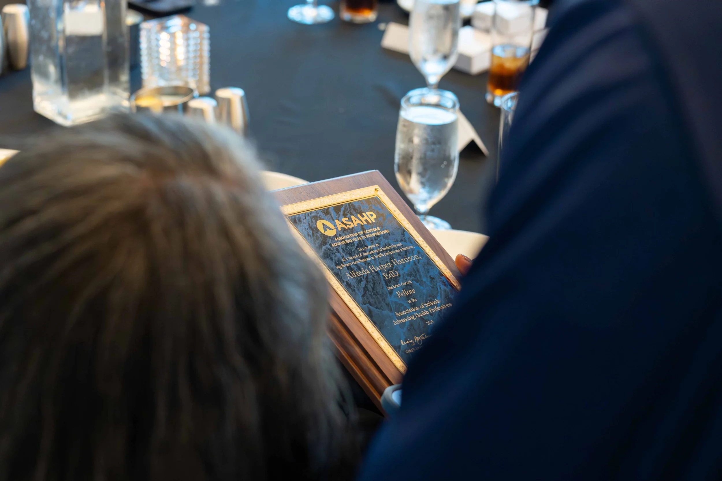 Close-up of a person reading an award plaque at a formal event with glasses of water, soda, and a black tablecloth visible.
