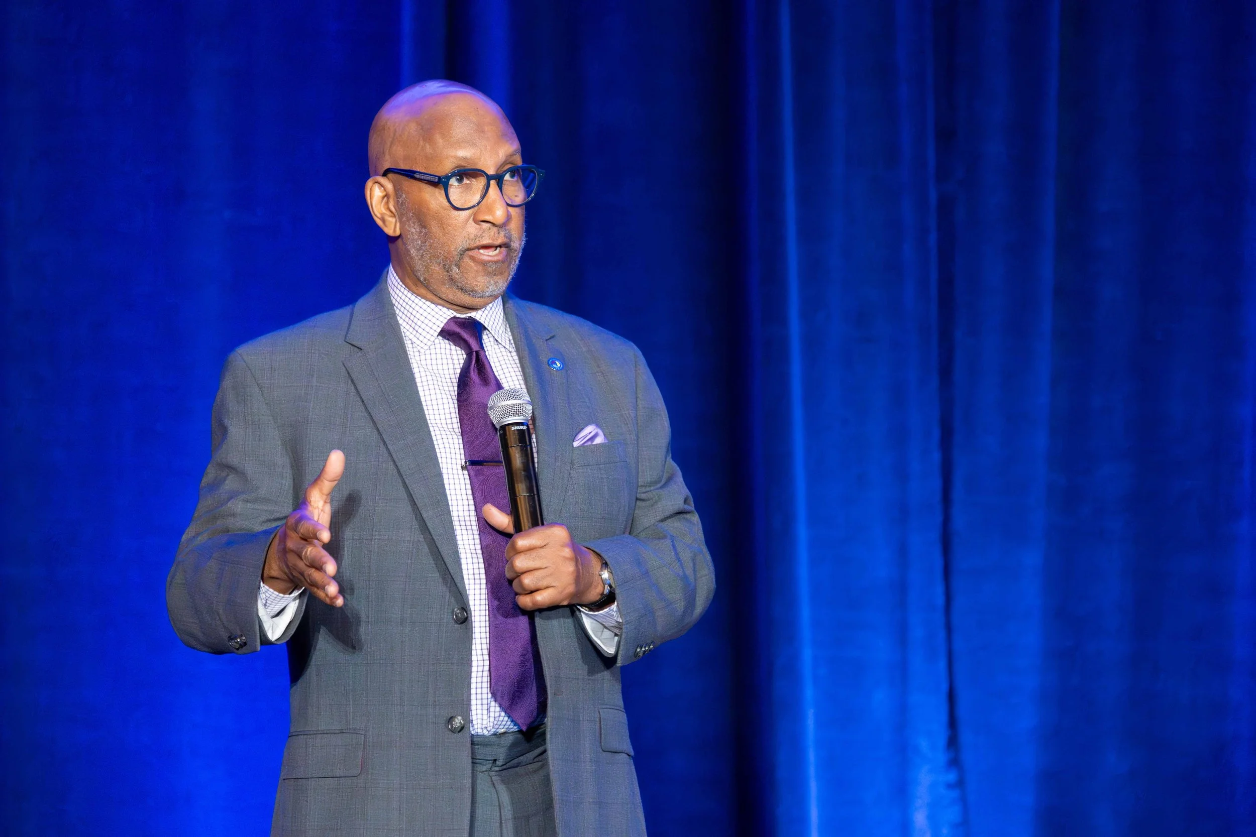A man in a gray suit, purple tie, and glasses speaking into a microphone on a stage with blue curtains.