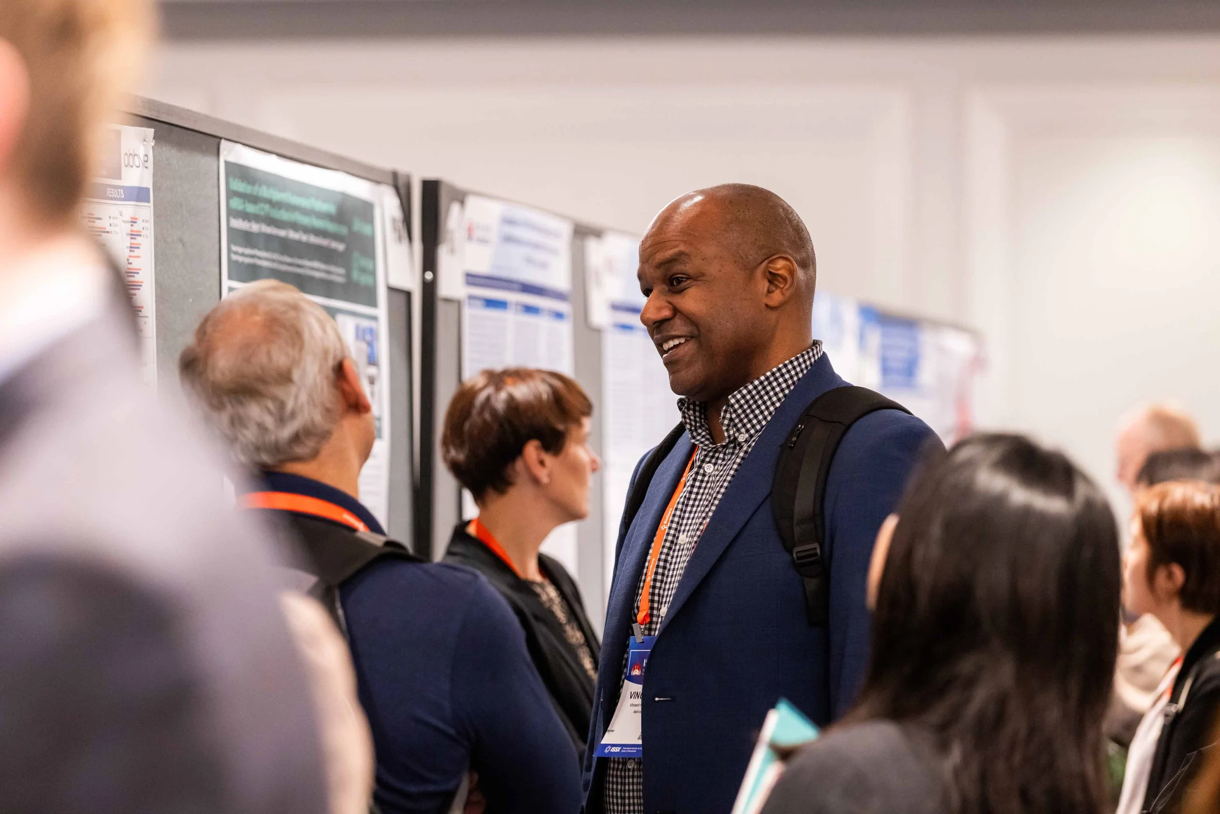 Man in a suit and backpack smiling and talking to a group of people at a conference or professional event with research posters on the wall.