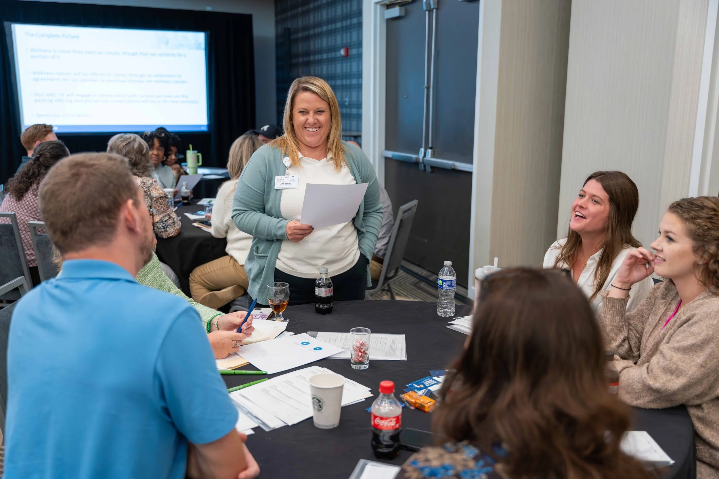 A group of people sitting around a conference table in a meeting room, with a woman standing and speaking to them while holding papers, others are smiling and engaging in conversation.