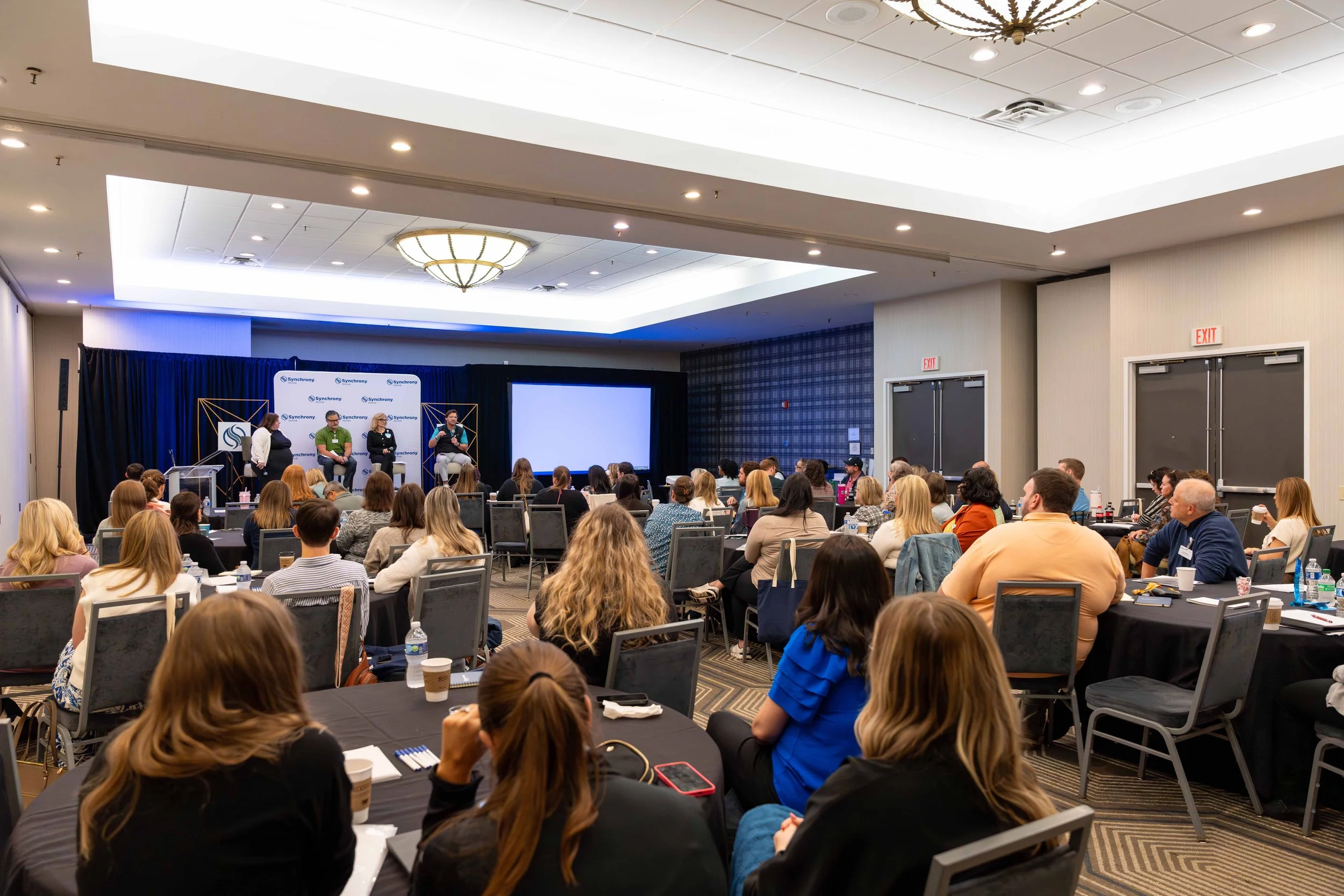Attendees listening to a panel discussion at a conference in a large meeting room with a stage, backdrop, and projection screen.