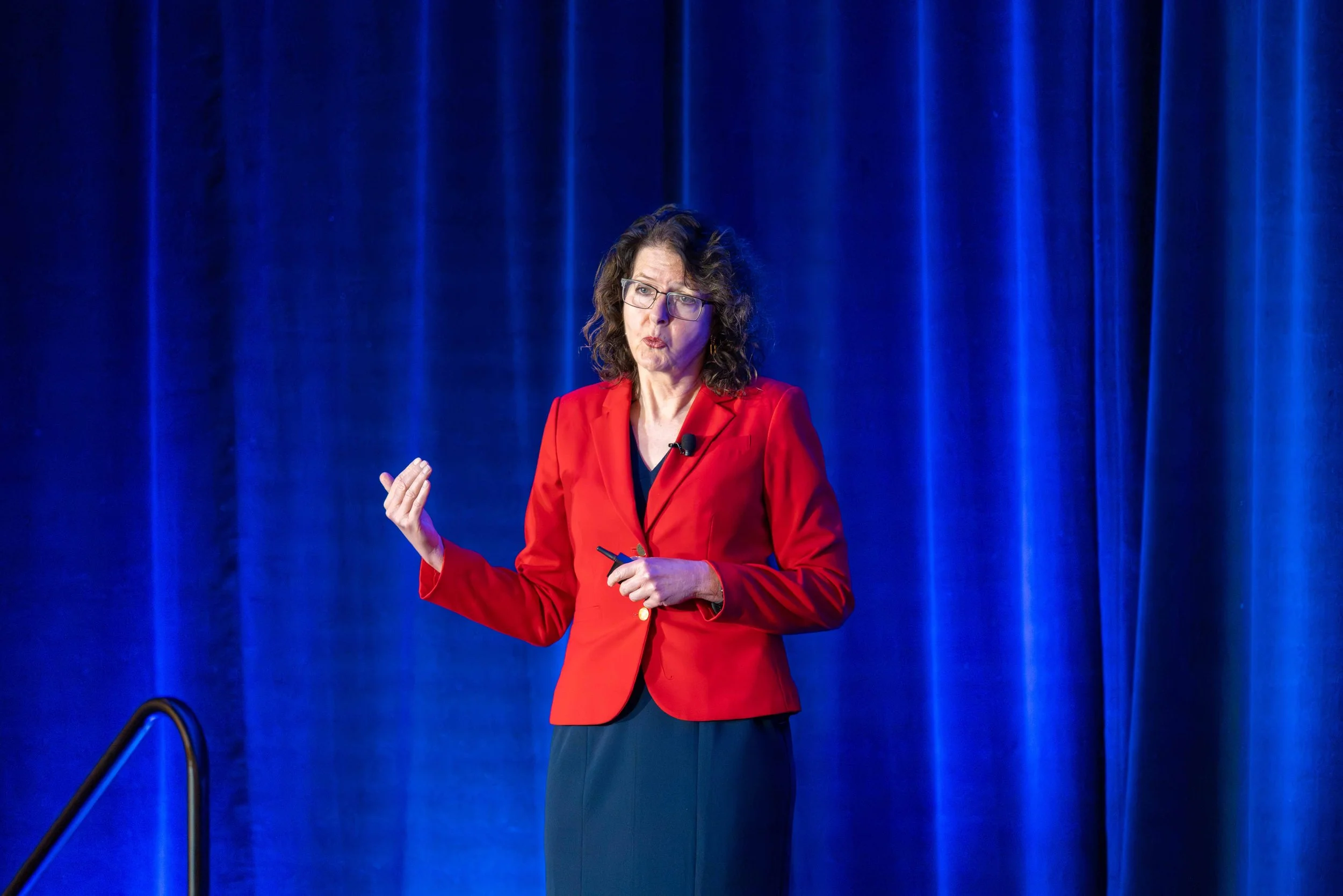 A woman in a red blazer giving a presentation in front of a blue curtain.