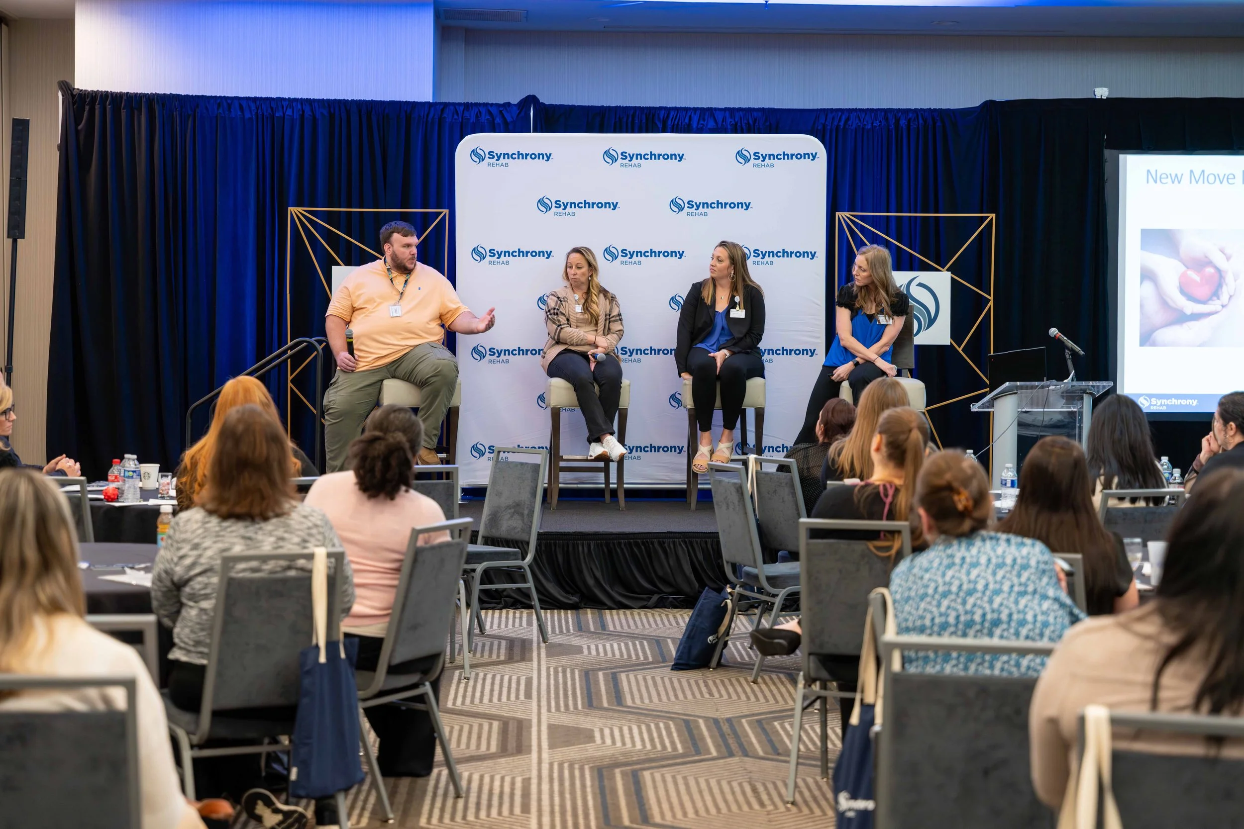 A panel discussion at a conference with four women and one man on stage, branded with 'Synchrony Rehab' banners, with an audience seated at round tables.