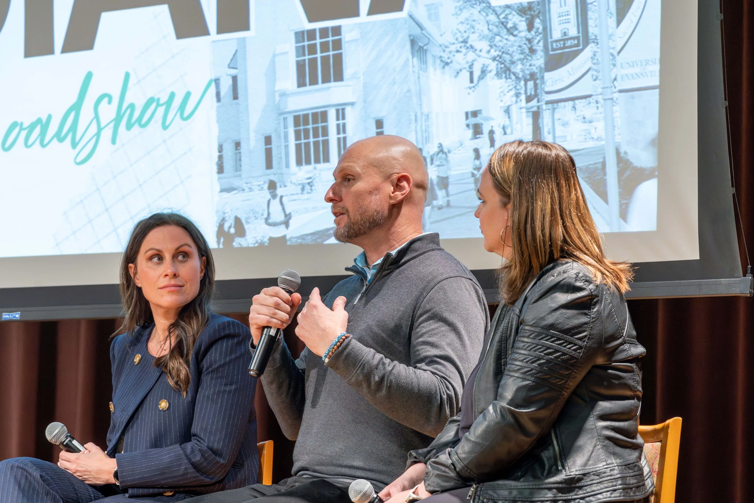 Three people speaking at a panel discussion, with a large screen behind them displaying the word "roadshow" and an outdoor campus scene.