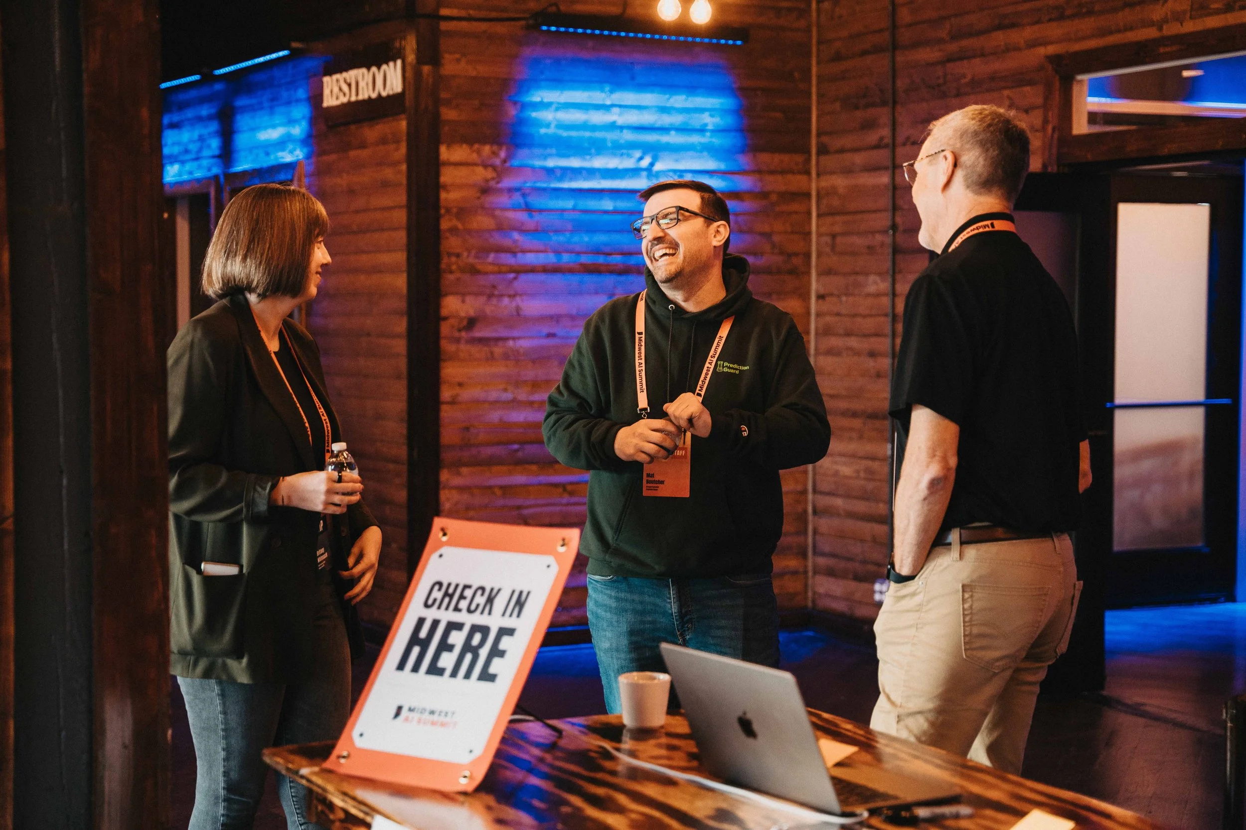 Three people are standing and talking near a check-in table with a laptop and a sign that says "Check In Here". The setting appears to be indoors with wooden walls and blue lighting. One person is holding a water bottle, and all are wearing lanyards.