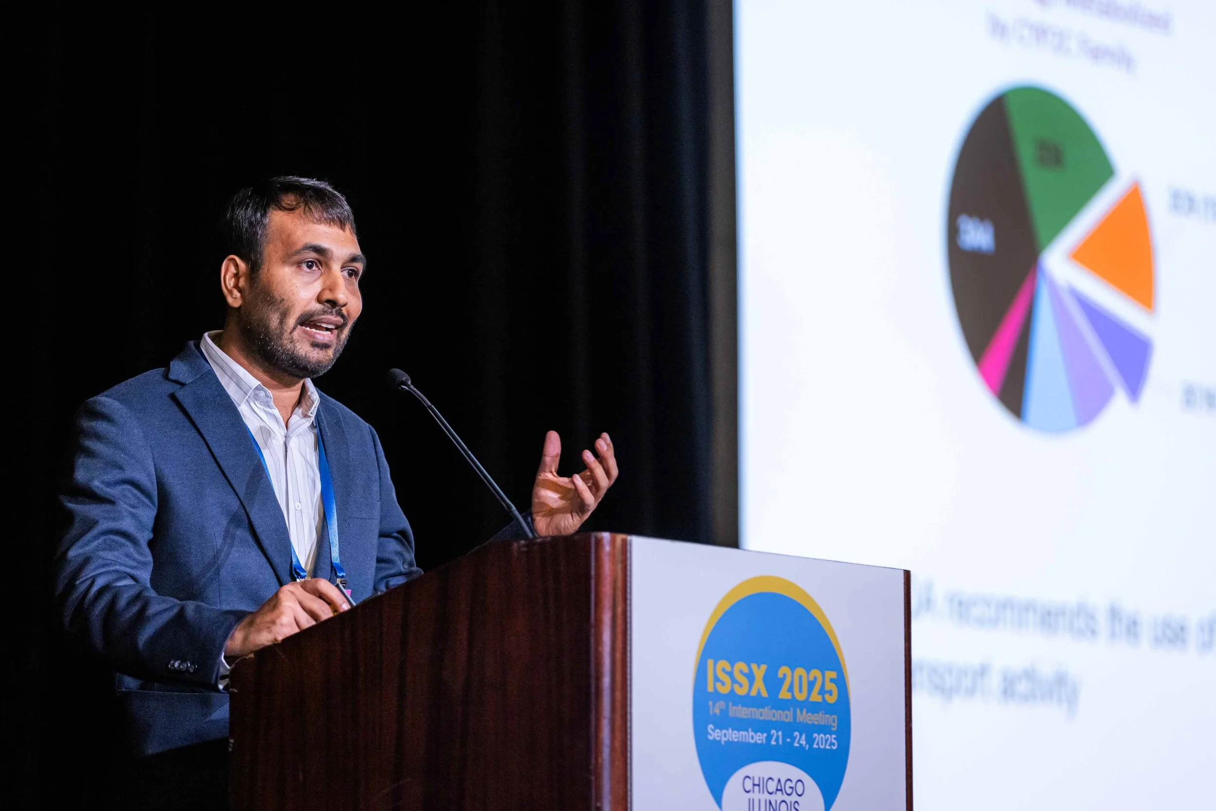 A man in a blue suit speaking at a podium during a conference, with a presentation slide showing a pie chart in the background.