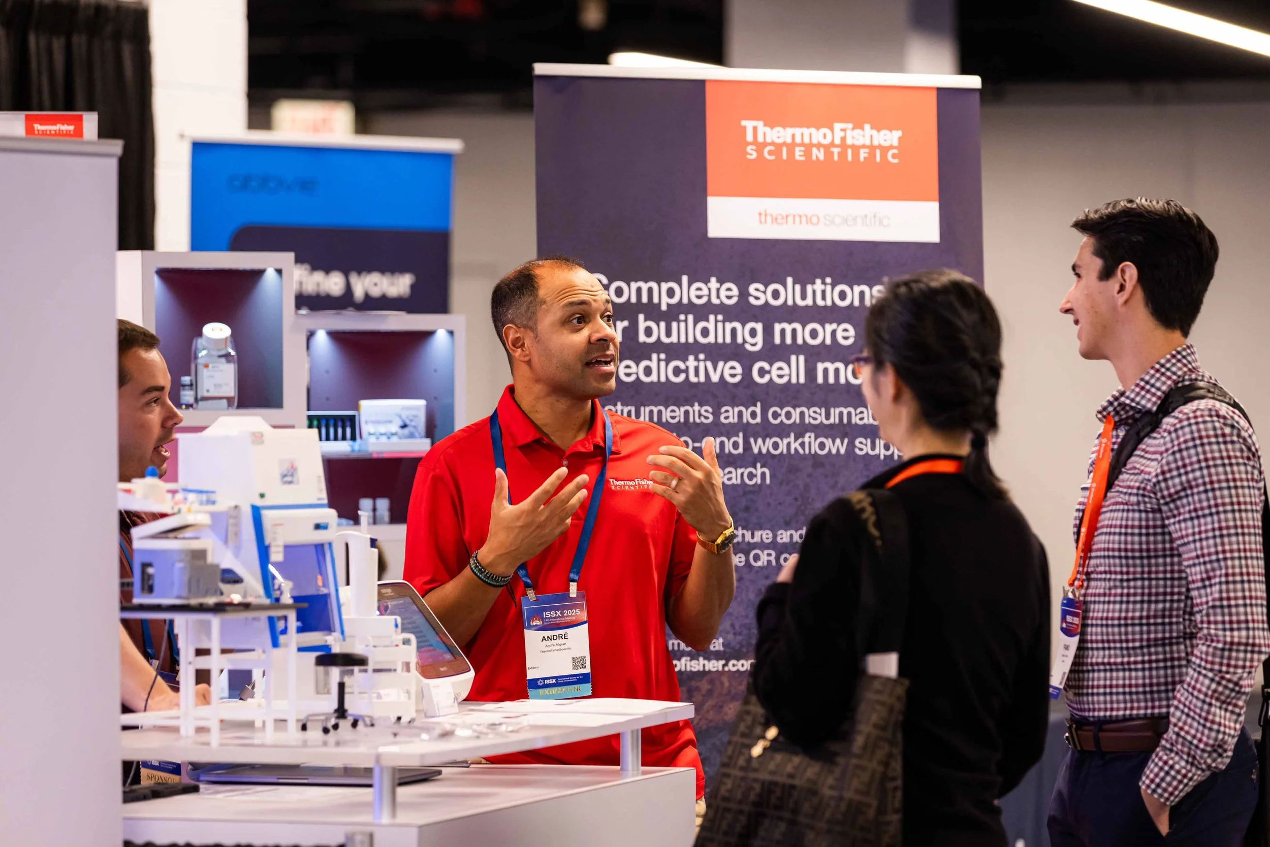 A group of four people at a trade show booth having a discussion. One man in a red shirt, wearing a name badge, is speaking to a man and two women who are listening attentively. The booth features a sign for Thermo Fisher Scientific and various scien