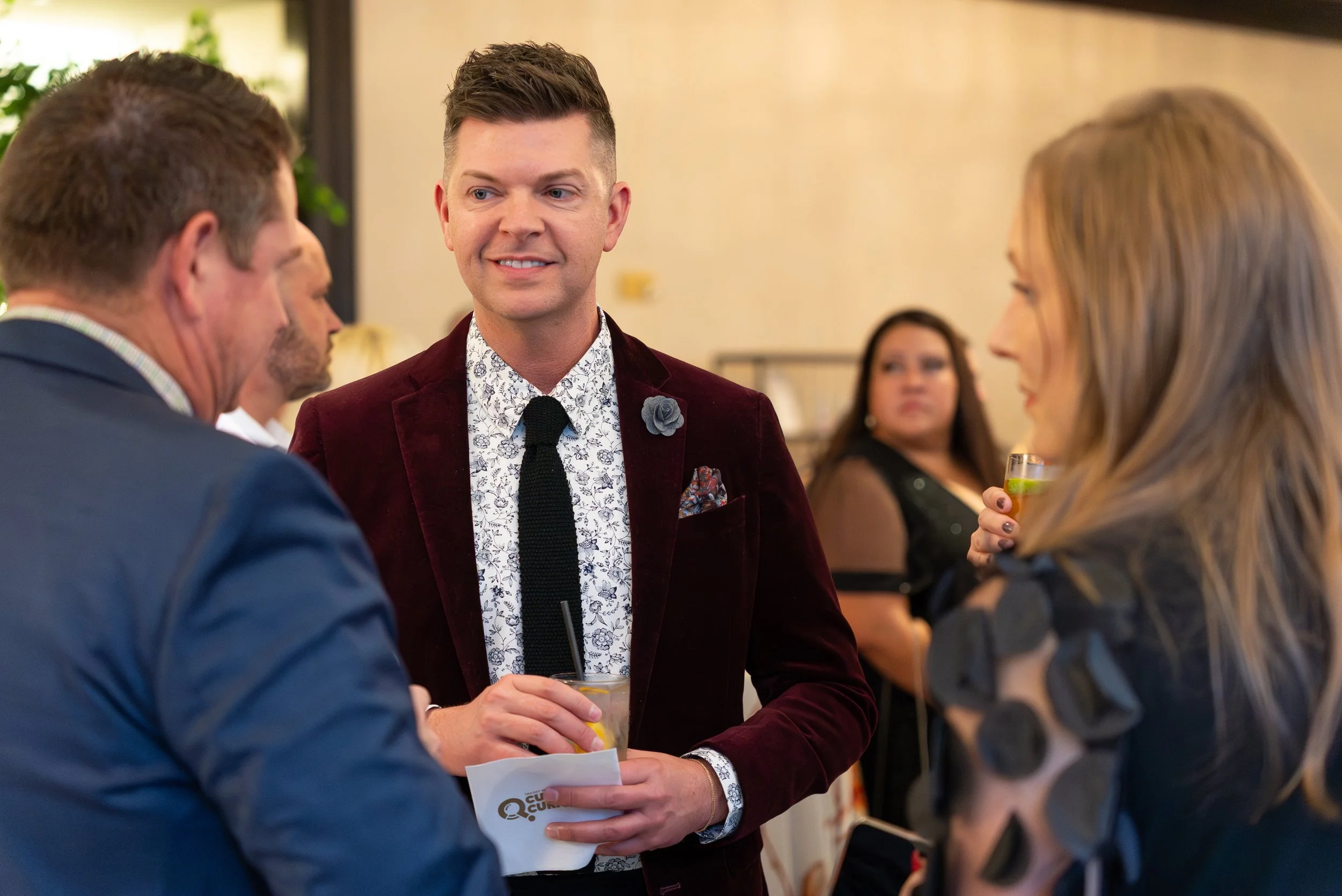 A man in a burgundy blazer, white patterned shirt, and black tie standing and conversing with a woman with blonde hair holding a drink, while another man in a blue suit also holds a drink nearby at a social gathering.
