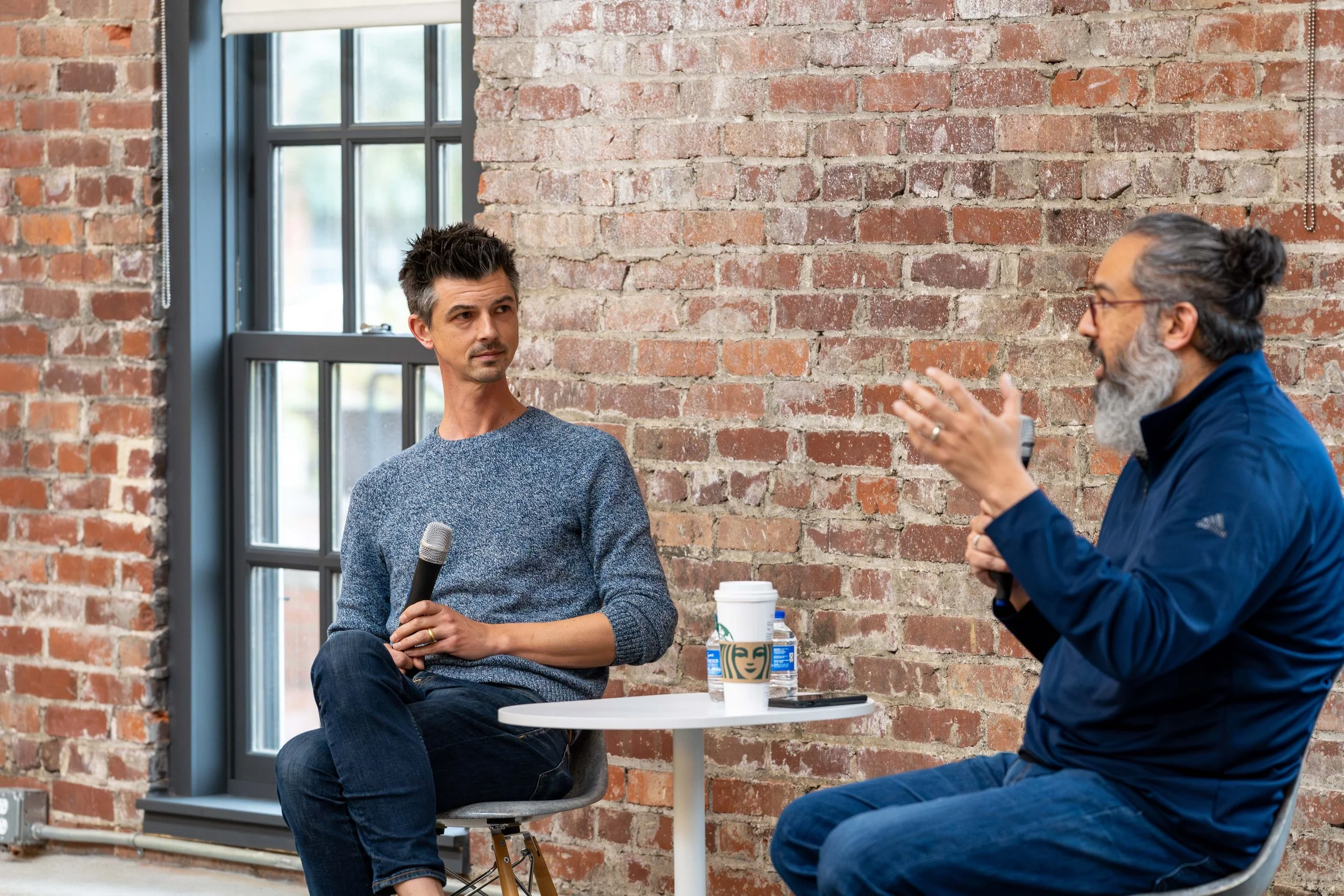 Two men sitting in a discussion, one with a microphone and the other gesturing, in front of a brick wall, with coffee cups on a small round table between them.