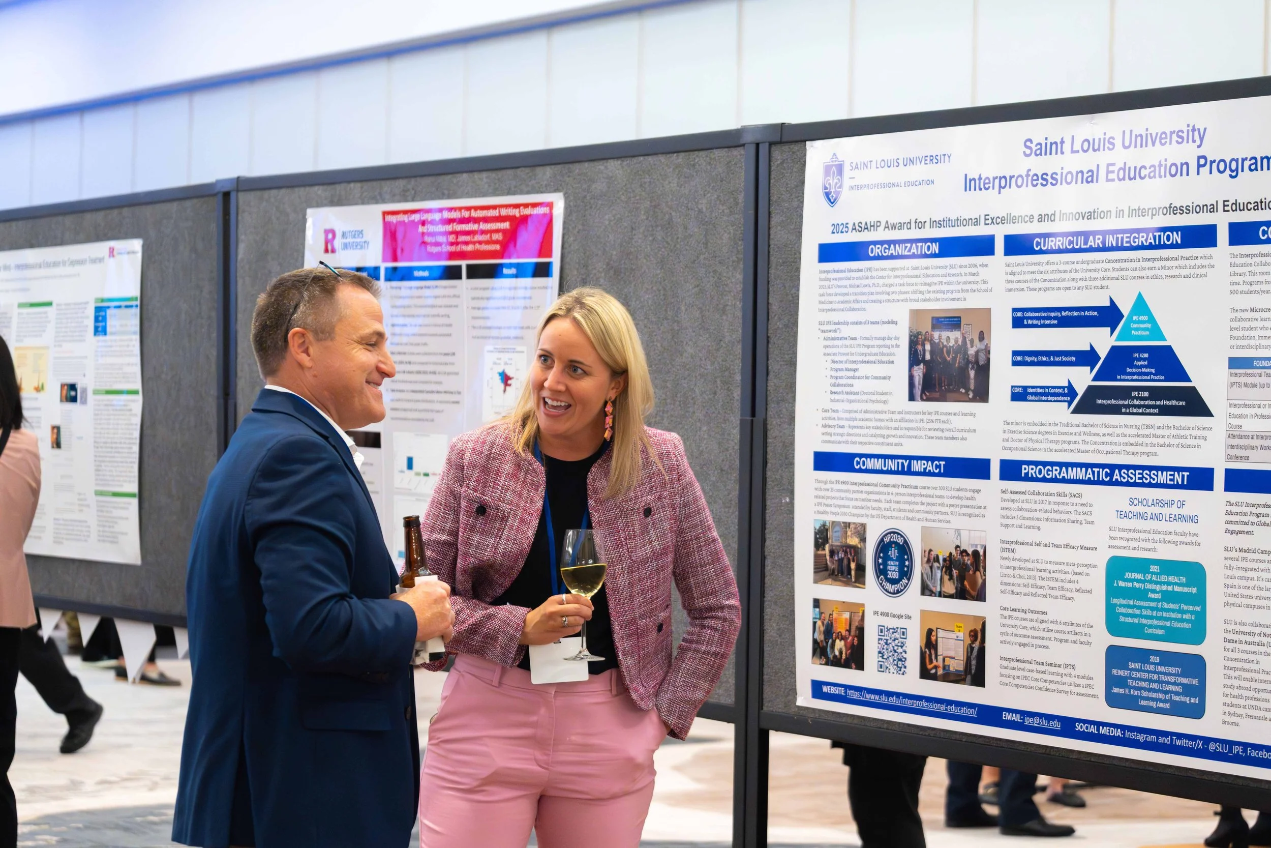 Two people, a man in a blue suit and a woman in a pink blazer, smiling and holding drinks, engaging in conversation at a professional conference with presentation posters on display behind them.