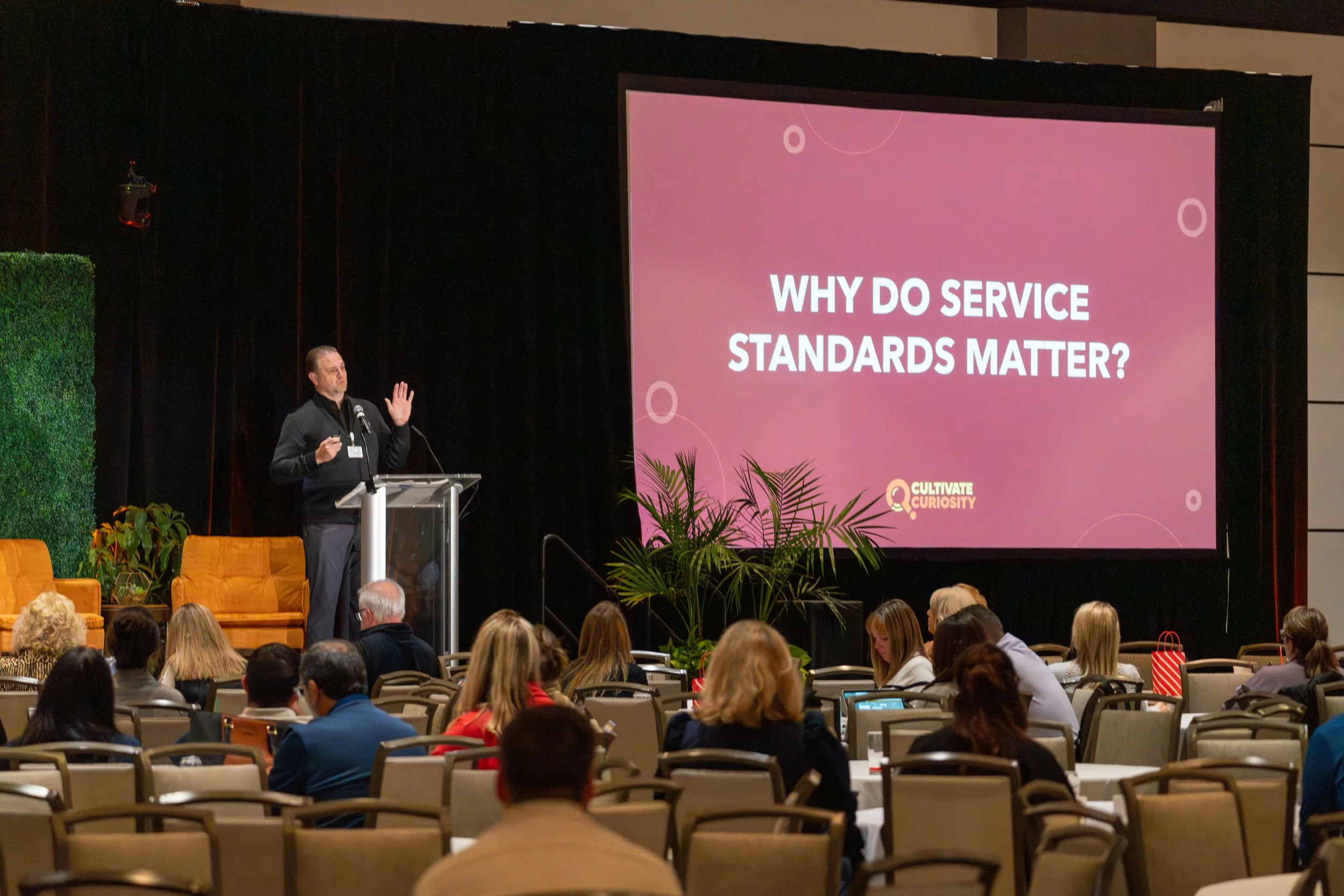 A man giving a presentation on stage at a conference, standing at a podium with a large screen behind him that reads, 'Why do service standards matter?' Audience members seated in chairs listen, some taking notes, in a conference room with plants and