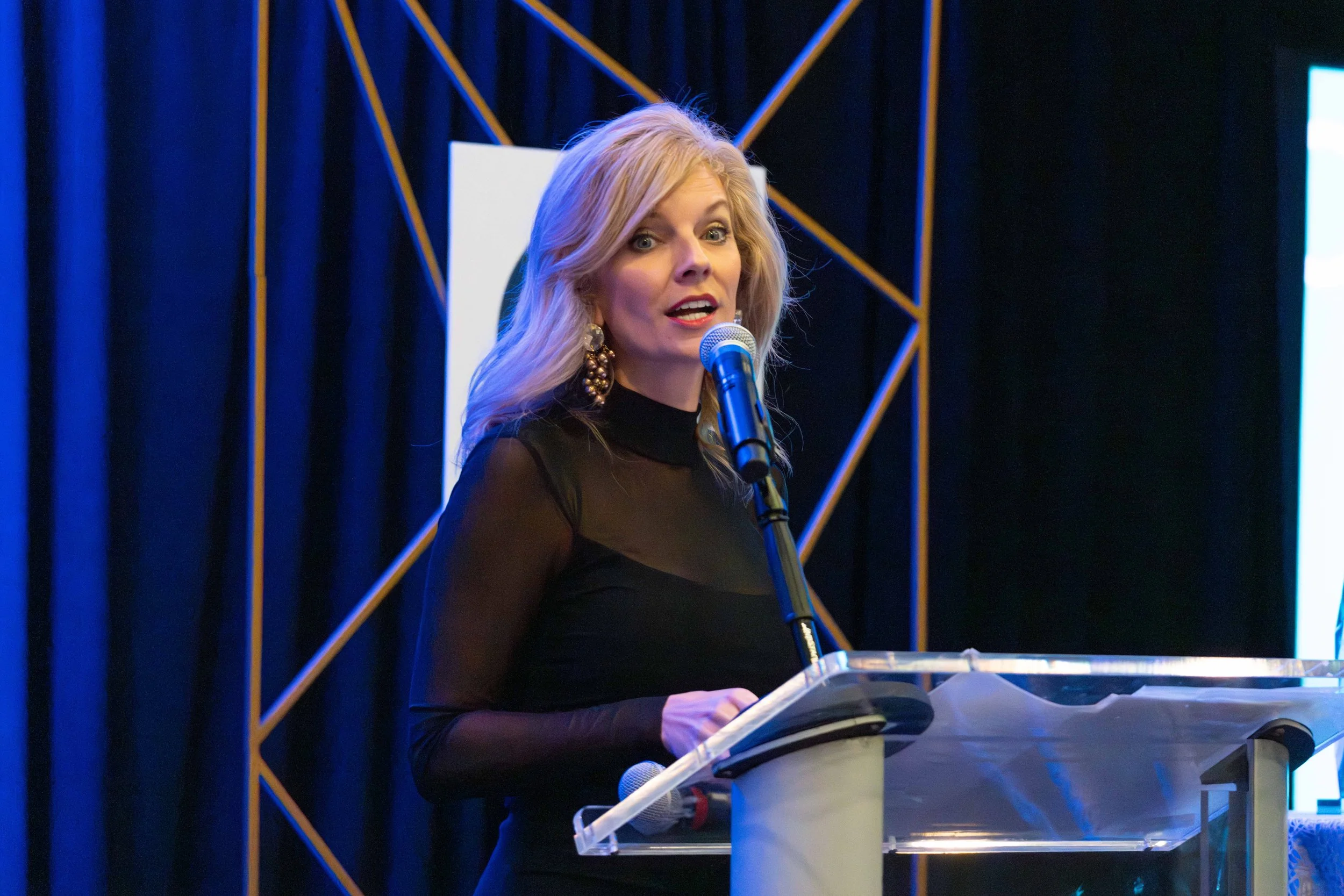 A woman with blonde hair wearing a black sheer top and large earrings speaking into a microphone at a podium during a formal event.