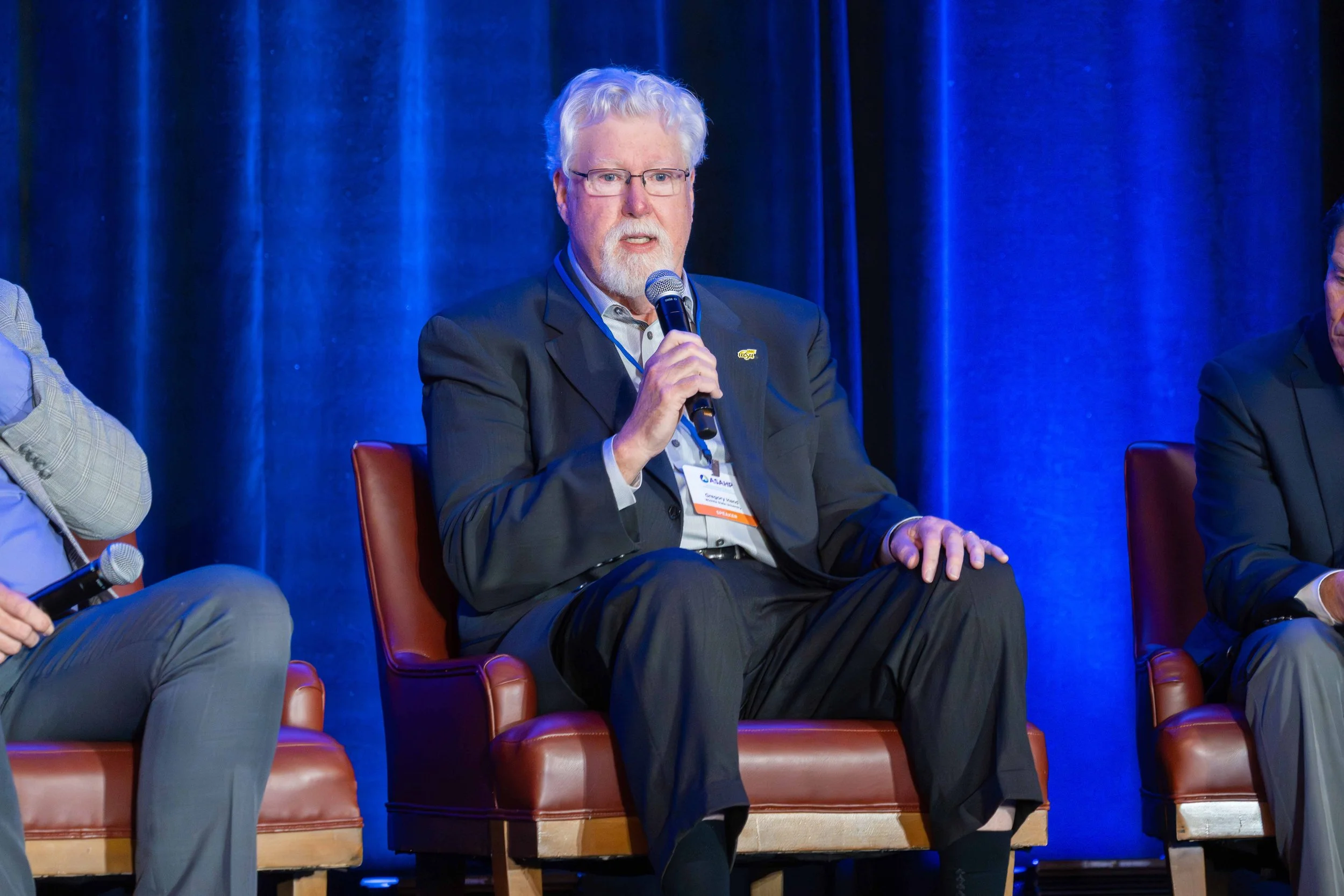 A man with white hair, glasses, and a beard sitting on a stage, holding a microphone, wearing a dark suit, white shirt, and a lanyard. He appears to be speaking at a panel discussion with dark blue curtains in the background.