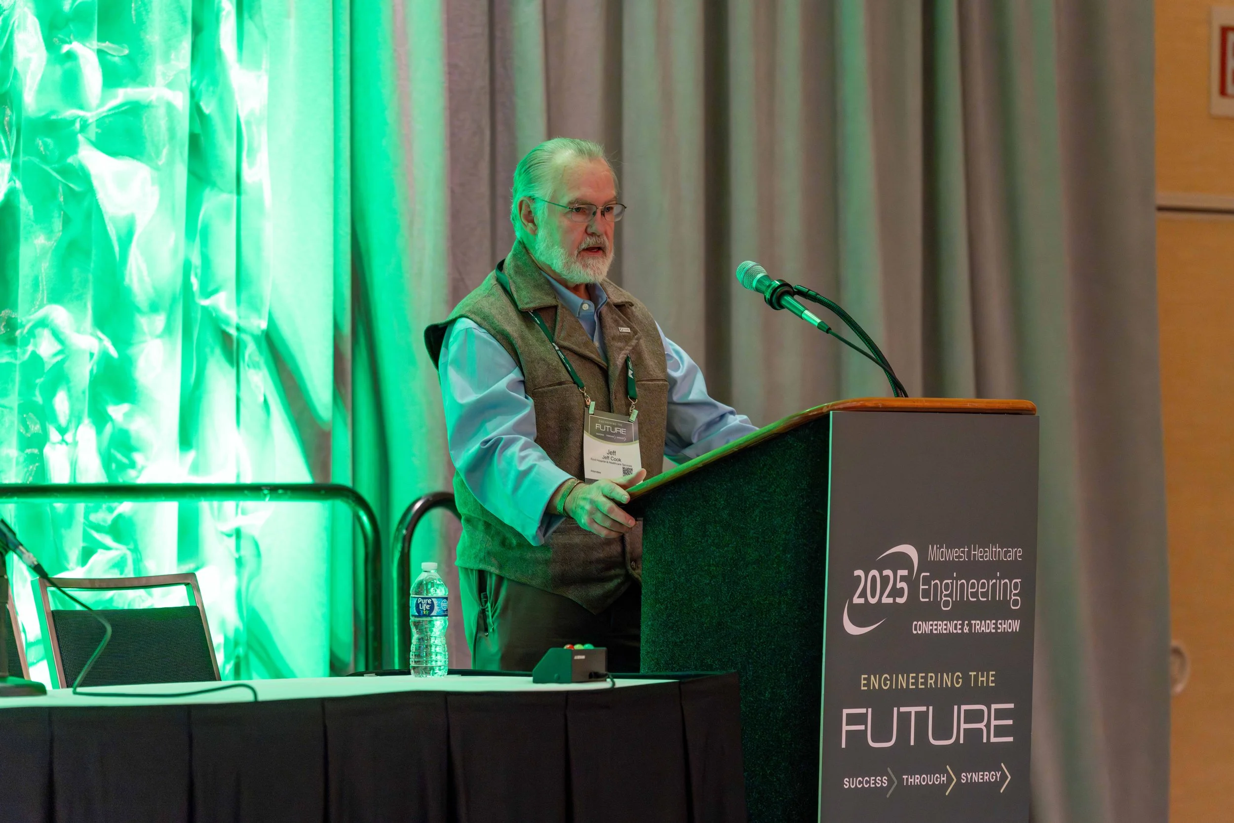 A man with gray hair and glasses standing at a podium giving a speech at a conference. The podium has a sign that reads 'Midwest Healthcare Engineering 2025 Conference & Trade Show' and 'Engineering the Future'. There is a water bottle and a chair on