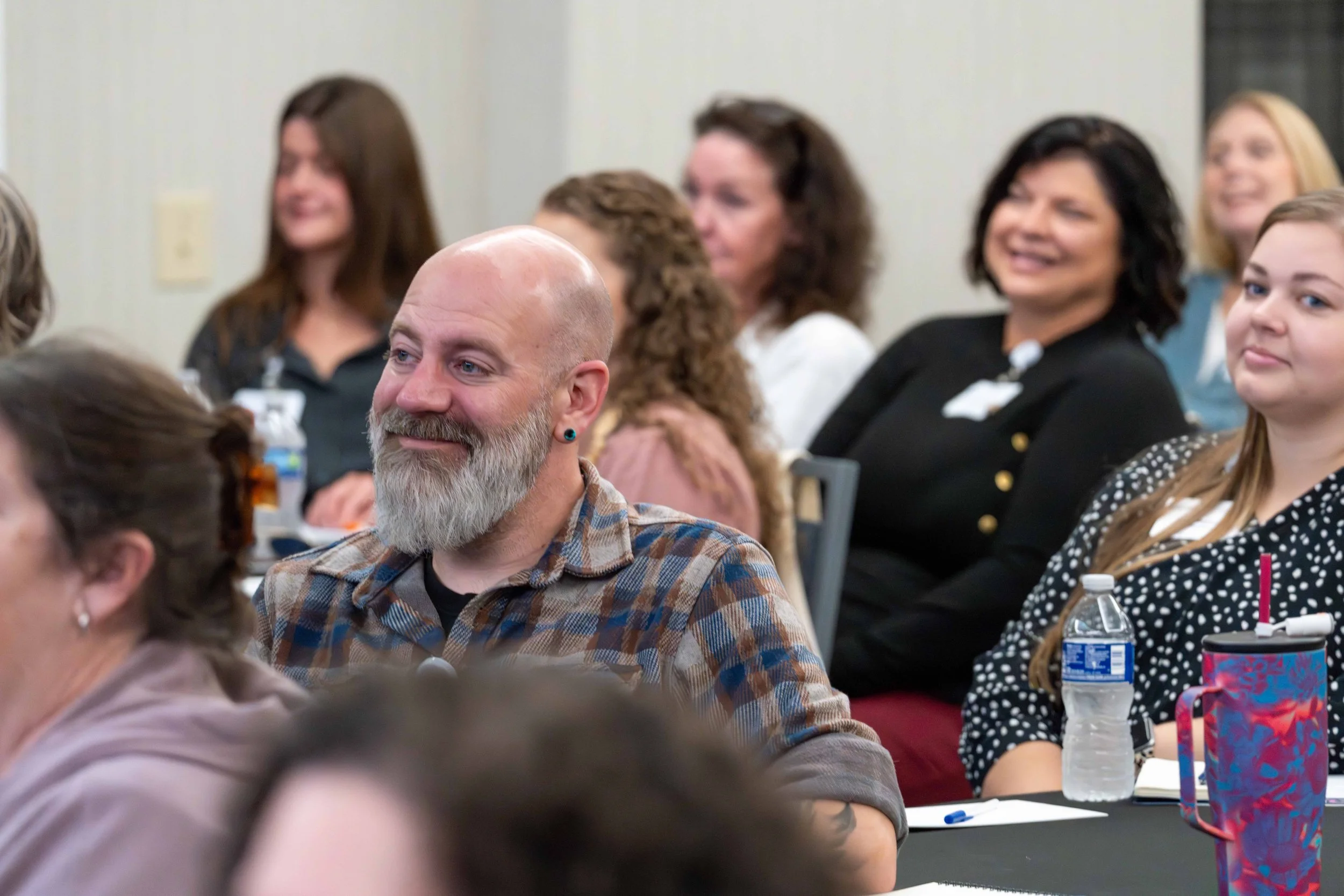 People attending a conference or seminar, sitting at tables, listening attentively, with some smiling and engaged.